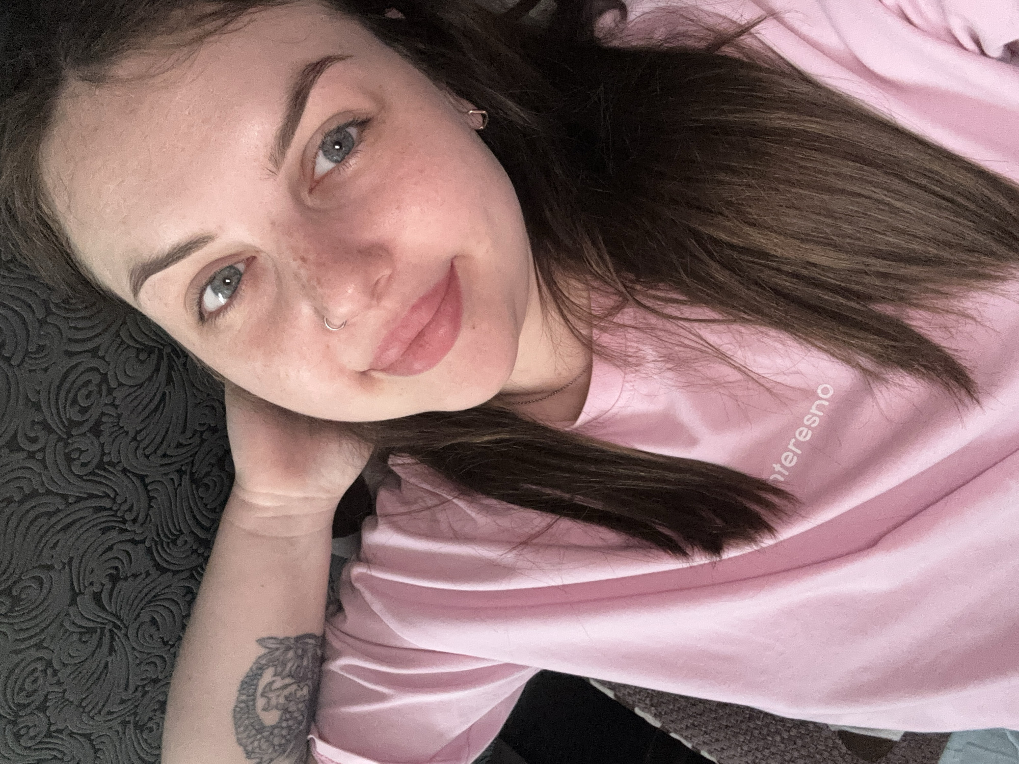 A woman with fair skin, freckles, and light blue eyes is lying down, looking up at the camera. She has dark brown hair that is long and straight. She is wearing a light pink t-shirt with a word visible on the shirt. She has a silver hoop in her nose and a silver earring. Her left arm is visible with a tattoo on it. The background shows a gray patterned surface next to her face, and clothing beneath her. There is no evidence suggesting that the photo was taken in Chornomorskoe, Ukraine.