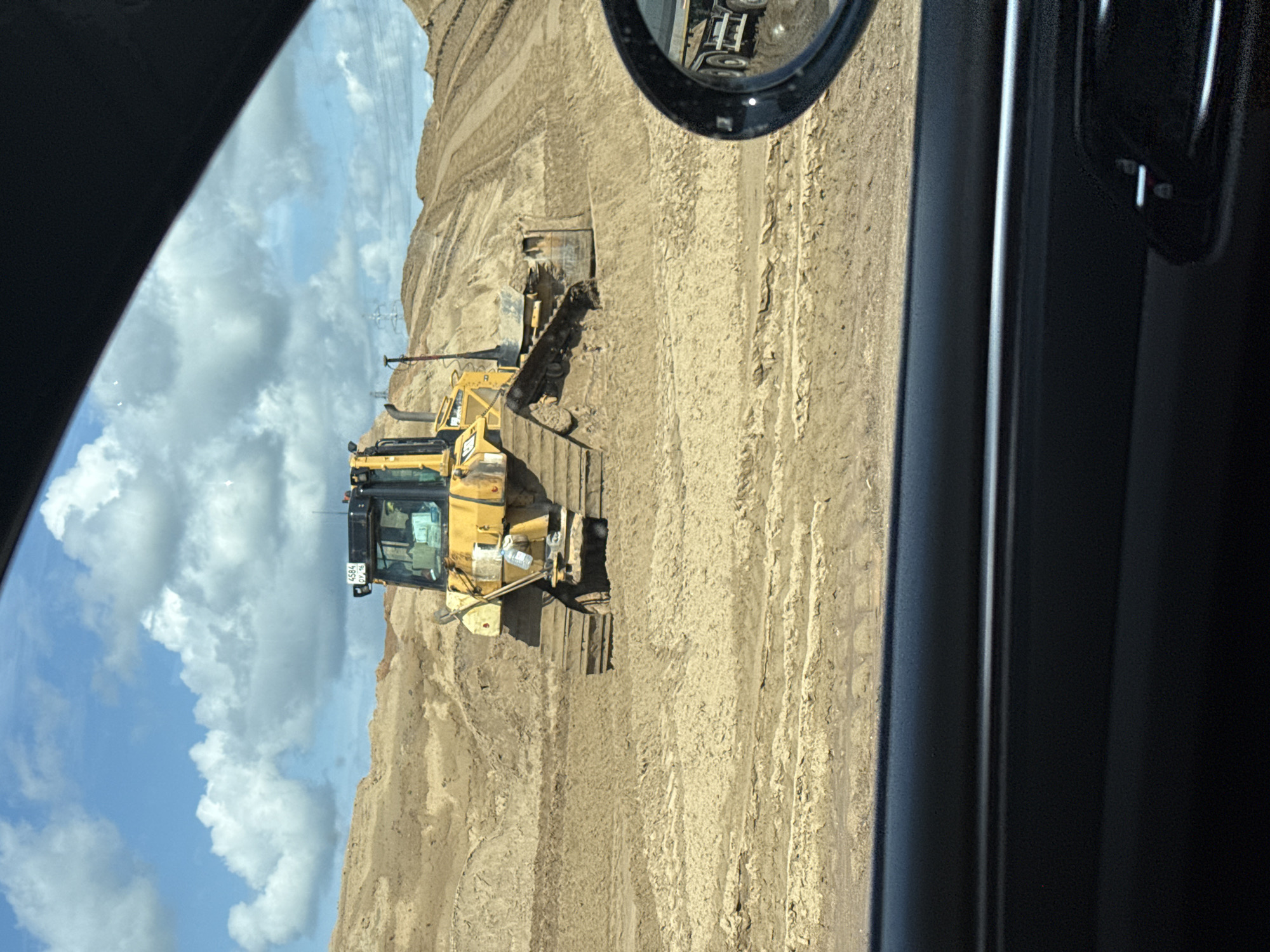 A yellow bulldozer operates in a muddy, sandy construction site under a cloudy sky. The bulldozer is the central focus, positioned on tracks partially submerged in mud. In the foreground, a car's side mirror reflects a trailer truck. The background shows a vast landscape of sand mounds, indicative of a large-scale excavation or construction project. The scene appears to be in Kabanikha, Russia, suggesting a construction site or quarry operation in the region.