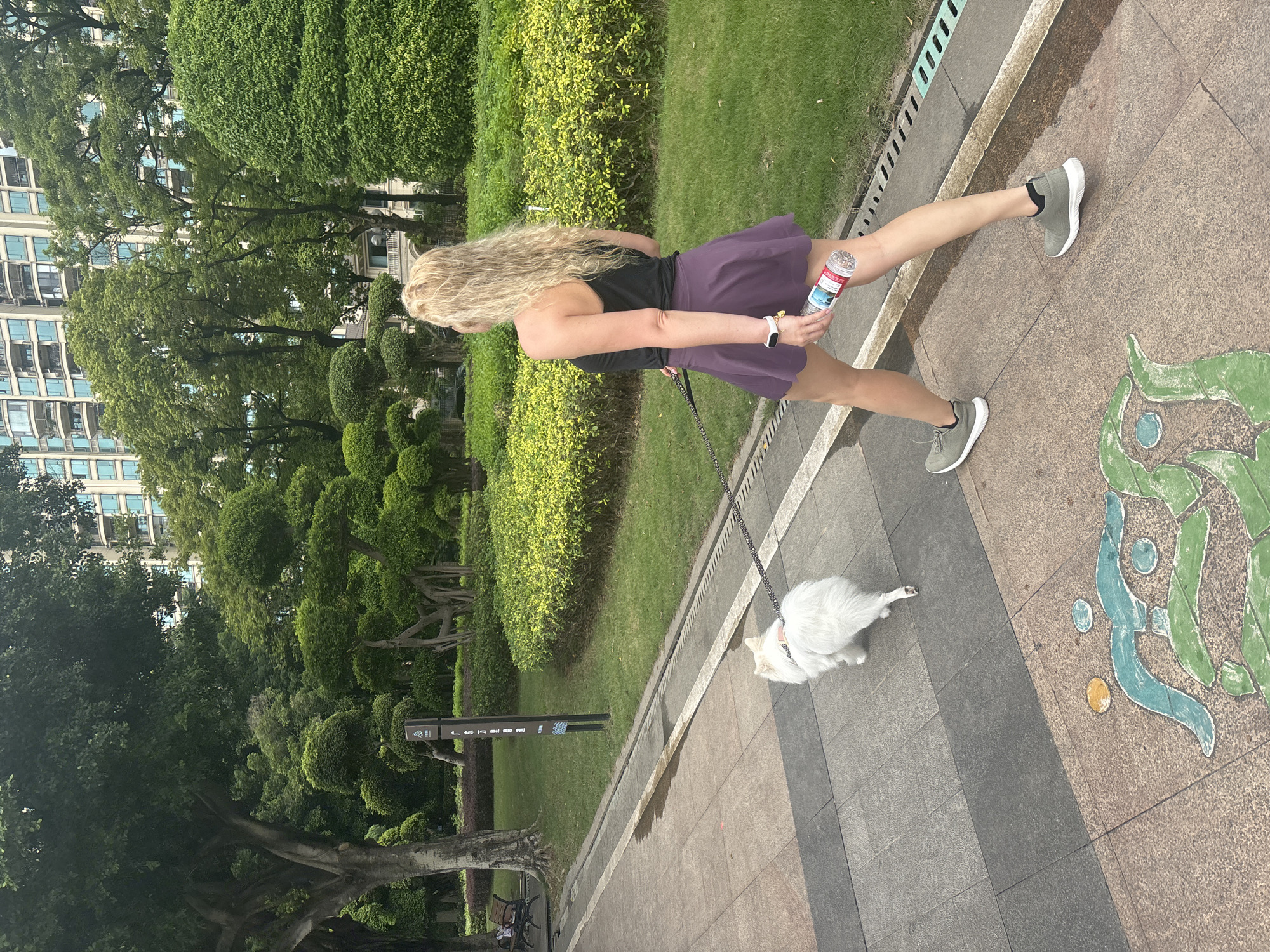 In the city of Nanhai District, China, a blonde woman with long, wavy hair, wearing a black tank top and a purple skirt, walks on a paved pathway. She is holding a clear plastic water bottle in her right hand and is connected by a leash to a small, white fluffy dog walking beside her. The pathway is bordered by a manicured lawn and features decorative colored patterns embedded in the pavement. In the background, trees with sculpted canopies and a tall, modern building are visible. The scene appears to be a park or public garden area.