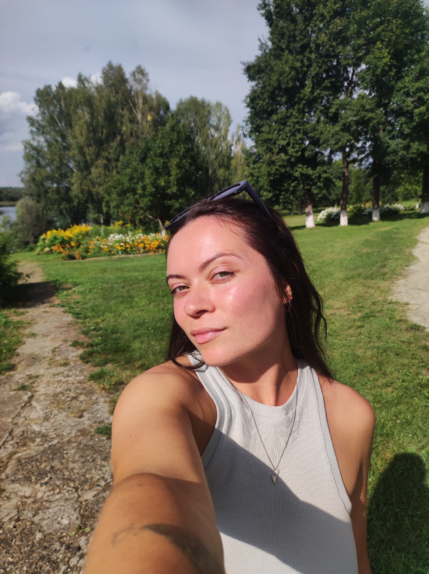 In Zhigalki, Russia, a young woman is taking a selfie outdoors in a park. She is positioned in the foreground, with her face and upper torso filling the right side of the frame. She has long, dark hair and is wearing a light grey tank top and a silver necklace. Sunglasses are perched on top of her head. The background features a paved path on the left, a lush green lawn, and a vibrant flower bed with yellow and orange flowers. Tall trees line the horizon, and a body of water is visible in the distance. The lighting suggests a sunny day.
