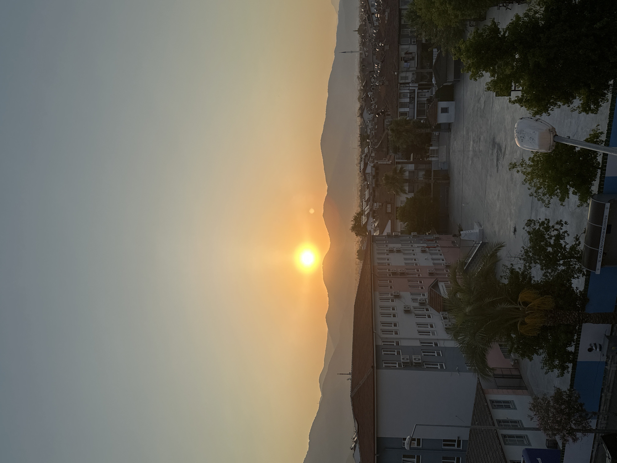 An outdoor, high-angle, full-shot captures a hazy, golden sunset over the city of Fethiye, Turkey. The sun, a bright, luminous circle, is positioned slightly left of center in the upper half of the frame, casting a warm glow that illuminates the sky and the distant mountains. The sky transitions from a pale blue at the top to a soft orange and yellow near the horizon.
In the foreground, a large, multi-story school building with a reddish-brown tiled roof dominates the left side of the image. It is painted in alternating bands of light blue and white, with numerous windows and what appear to be air conditioning units mounted on the facade. To the left of the school is a paved courtyard, empty except for a tall, modern lamppost with a white fixture.
A tall palm tree with yellowing fronds stands near the center of the courtyard, partially obscuring the school building. To the right of the courtyard, a densely packed residential area with terracotta-tiled roofs stretches towards the mountains. A minaret of a mosque is visible rising above the rooftops in the distance.
The mountains in the background are silhouetted against the bright sky, softened by the atmospheric haze. Green trees are scattered throughout the scene, particularly in the mid-ground and foreground. The overall impression is of a peaceful, early evening cityscape under a dramatic sunset.