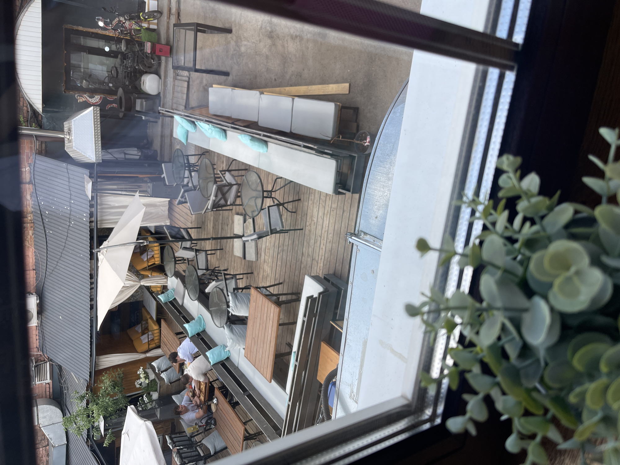 The image shows an outdoor patio viewed through a window. The patio has seating areas with tables and chairs, some covered by white umbrellas. There are people seated at some of the tables. There is also a section with bench seating and cushions. Various objects are stacked on the side near the window. A building with a metal roof is visible in the background. The image does not contain sufficient information to identify a specific location in Izhevsk, Russia.