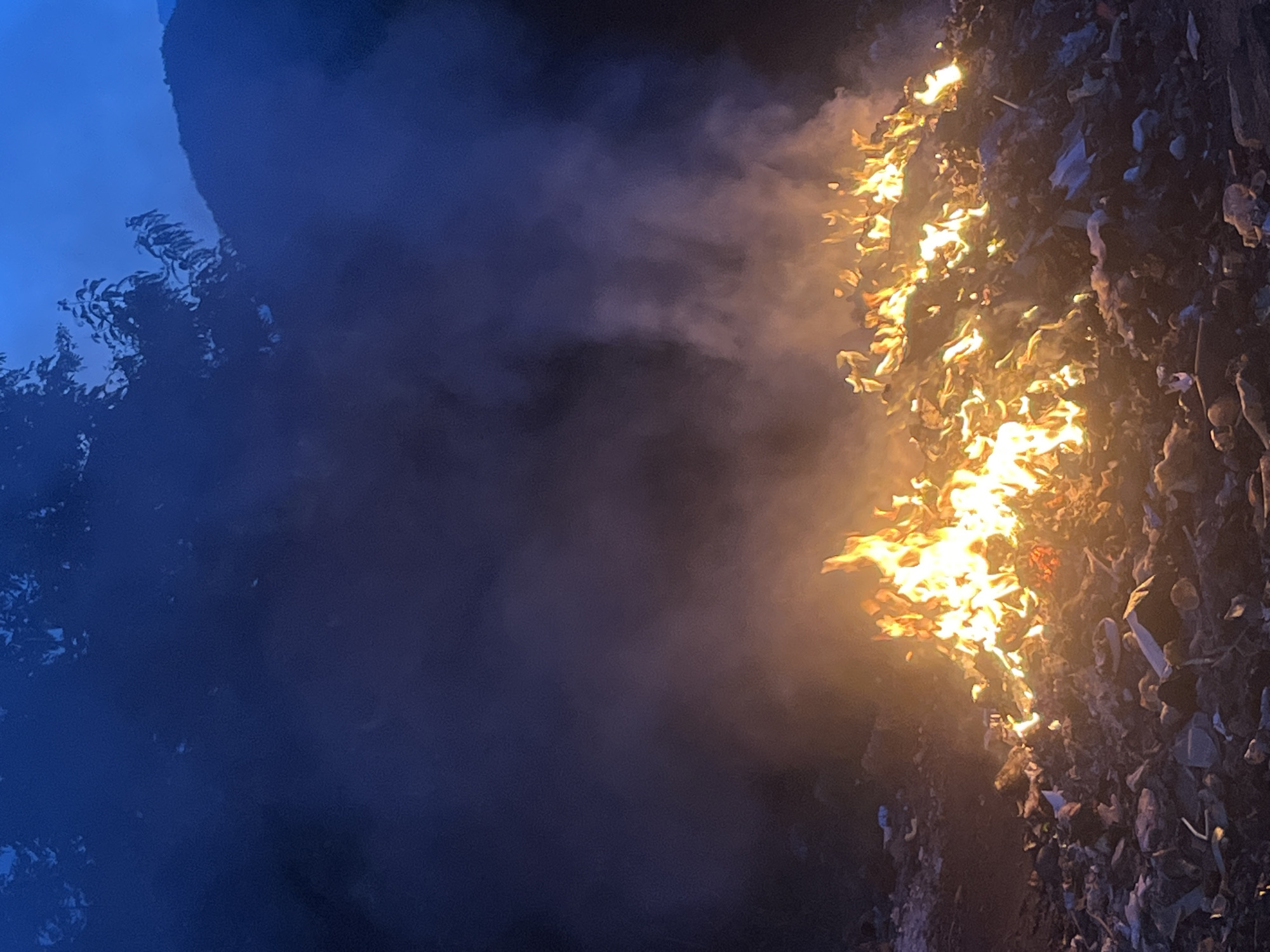 A burning pile of mixed refuse is seen at night, emitting smoke. The fire consumes the top portion of the pile, with unburnt debris below. To the left is a dark area that could be trees. The location is Bac Nha Trang Ward, Vietnam.