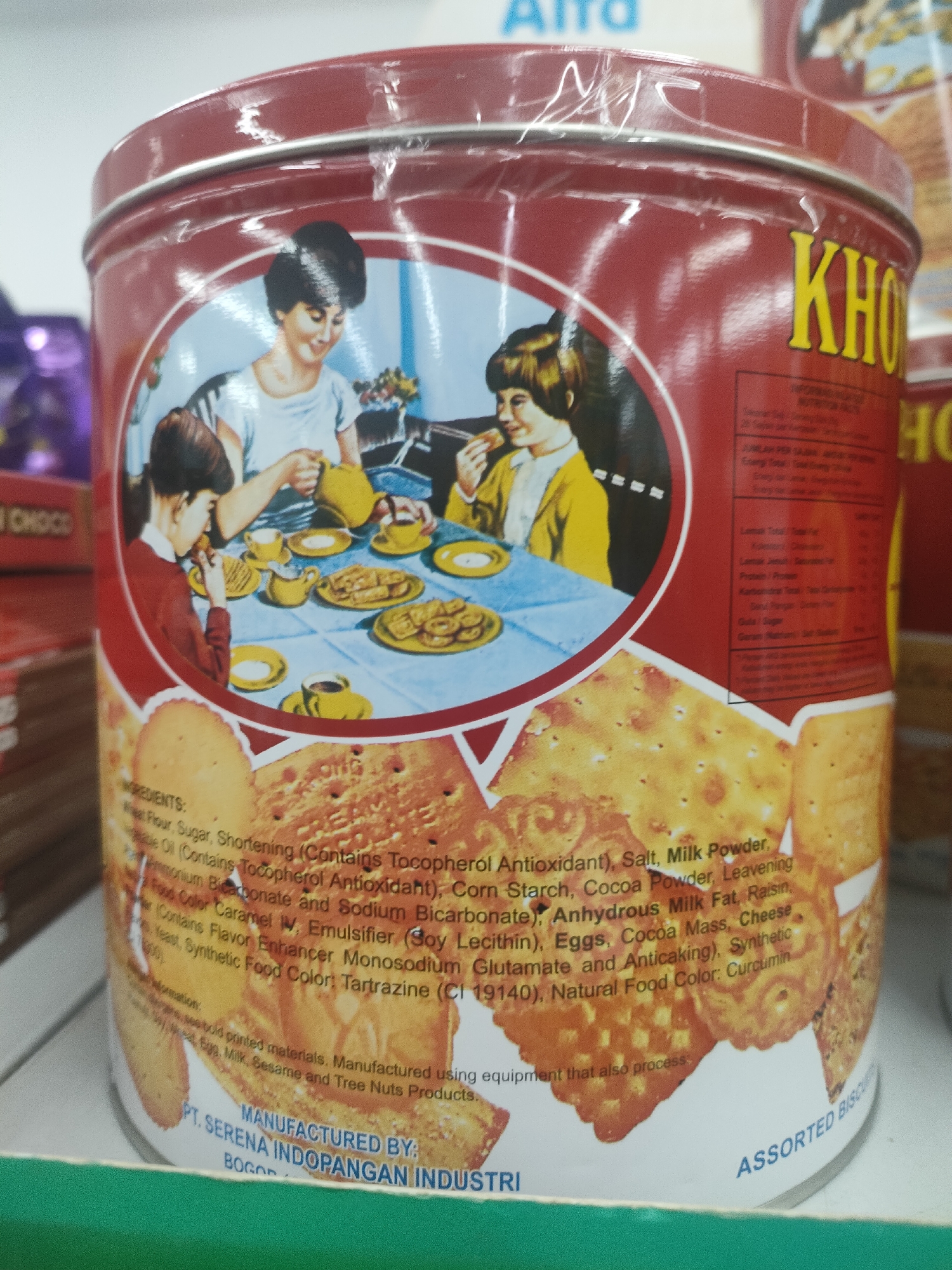 The image shows a close-up of a red tin can with an illustration of a family enjoying tea and biscuits. The illustration depicts a woman pouring tea for two children at a table laden with biscuits and cups. The can has text printed on it in various languages, including "KH" in large yellow letters on the right side. Below the illustration, a list of ingredients is partially visible, starting with "INGREDIENTS: Wheat Flour, Sugar, Shortening (Contains Tocopherol Antioxidant), Salt, Milk Powder, Vegetable Oil (Contains Tocopherol Antioxidant), Corn Starch, Cocoa Powder, Leavening Agents (Ammonium Bicarbonate and Sodium Bicarbonate), Anhydrous Milk Fat, Raisin, Cocoa Color Caramel IV, Emulsifier (Soy Lecithin), Eggs, Cocoa Mass, Cheese Flavor, Flavor Enhancer Monosodium Glutamate and Anticaking), Synthetic Sweetener, Synthetic Food Color: Tartrazine (CI 19140), Natural Food Color: Curcumin". Further down, it states "MANUFACTURED BY: PT. SERENA INDOPANGAN INDUSTRI BOGOR". The bottom of the can has "ASSORTED BISCUITS" printed on it. The background is blurred, showing what appear to be shelves and other products. The can is situated on a green surface. The overall impression is that of a product advertisement or display in a retail setting. The location "Desa Banyumas, Indonesia" is not depicted in the image.
