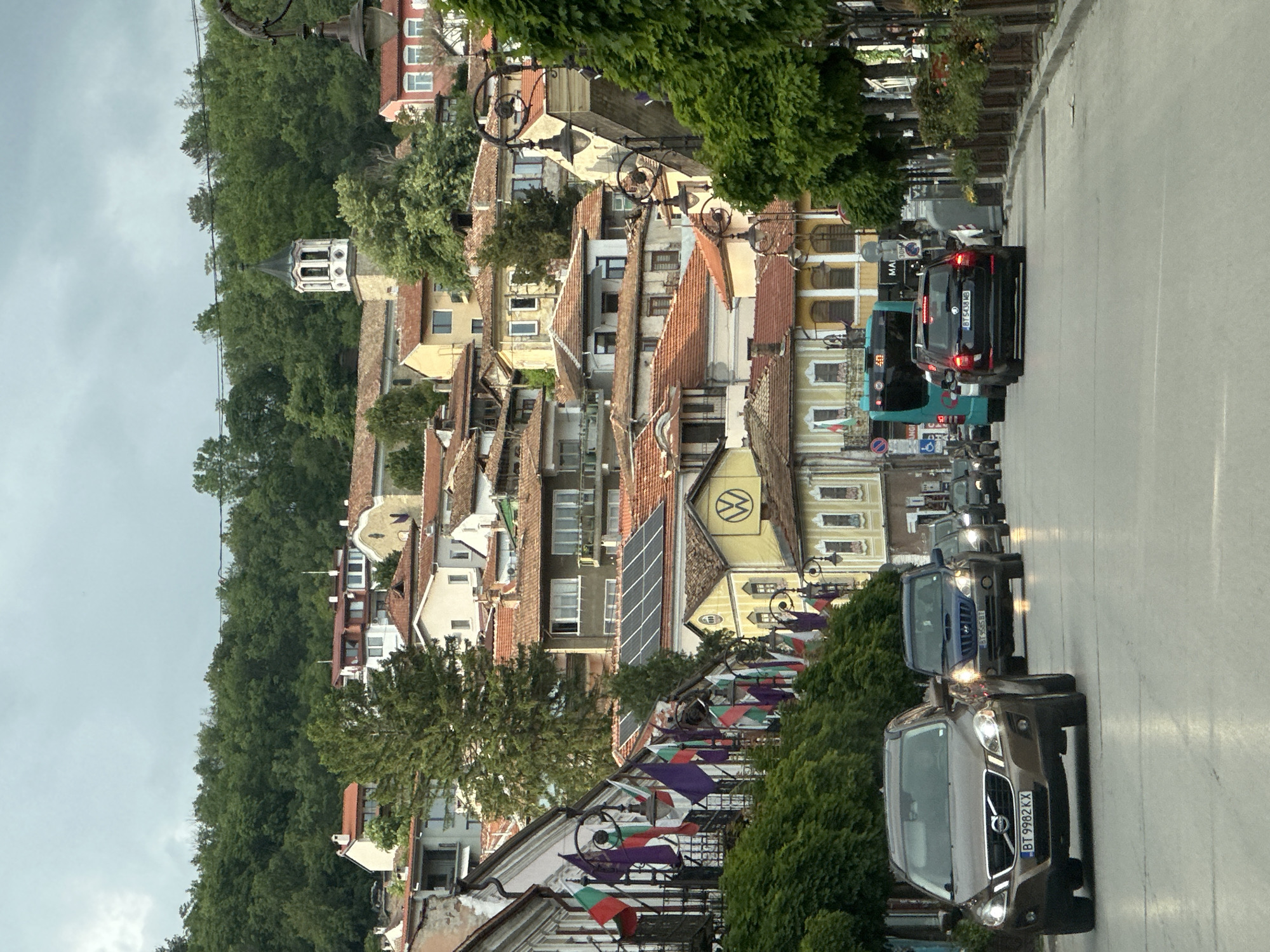 The image shows a street scene in Veliko Tarnovo, Bulgaria. The camera is positioned on a road lined with a mix of older buildings featuring terracotta roofs and newer constructions. The architecture reflects a blend of historical and modern styles. On one side of the road, the buildings are interspersed with dense greenery, including trees and bushes, contributing to a lush environment. Street lamps with decorative wrought iron designs line the road. Bulgarian flags are visibly displayed along the street.
The sky is overcast. The road itself shows moderate vehicular traffic, including cars and a bus, moving in the same direction. The lighting suggests it is daytime, but the weather conditions create a somewhat muted atmosphere. In the background, a tall, white church tower rises above the rooftops, punctuating the townscape.