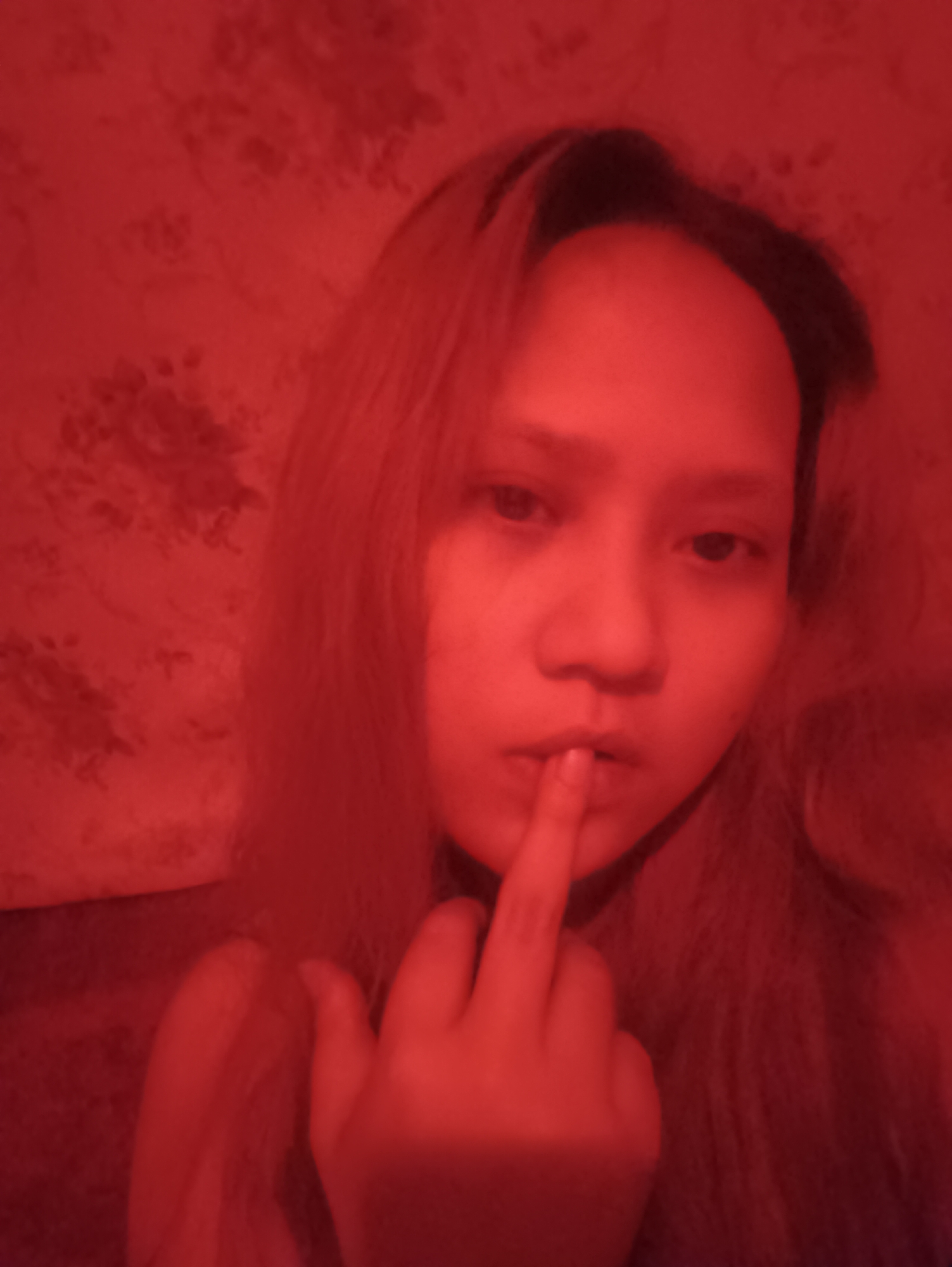 A person is captured in a close-up, selfie-style portrait, bathed in a strong red light that casts a reddish hue over the entire scene. The person's face is the central focus, with their right index finger placed against their lips, suggesting a gesture of silence or contemplation. Their hair, appearing to be light brown or blonde, is styled loosely around their face. The background features a red-patterned wallpaper, adding to the overall red ambiance of the image. The person's attire is not clearly discernible due to the lighting conditions. The image does not provide any specific details that would allow for its location to be identified in Depok, Indonesia.