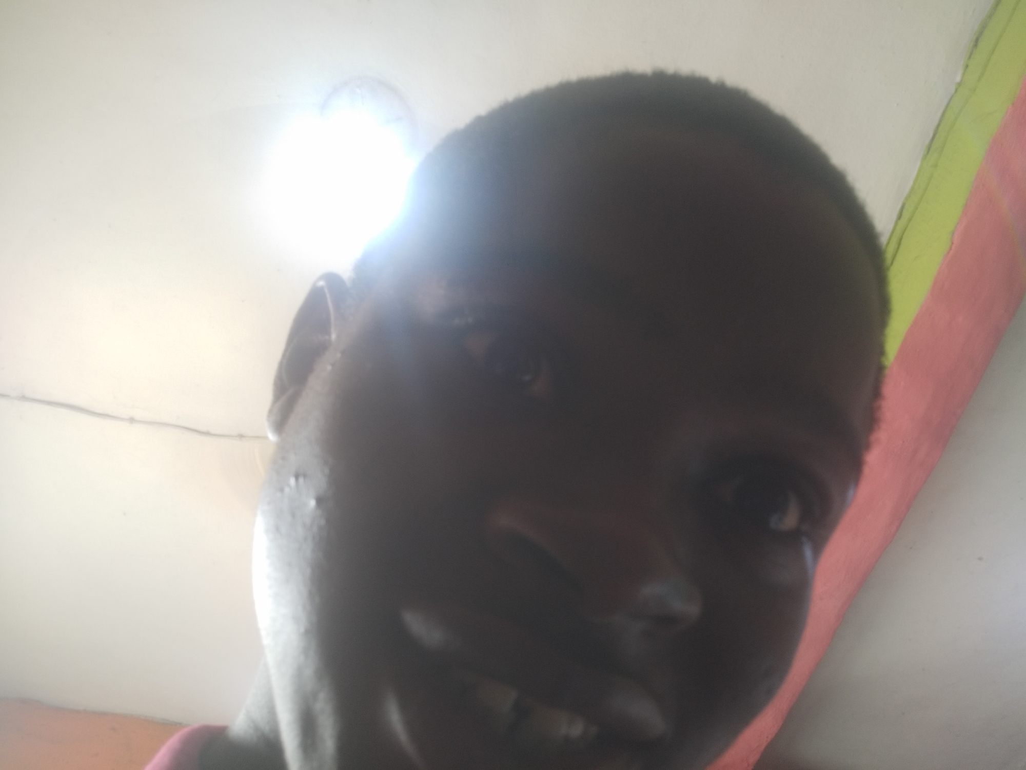 A close-up photograph shows a dark-skinned person, likely a male, with short hair. The person is smiling slightly, revealing teeth. The lighting is poor, with a bright light source positioned above and to the left, causing significant shadowing on the face. The background includes a white ceiling and a portion of a wall painted with horizontal stripes of red, green, and a lighter color. The photograph appears to have been taken indoors, possibly in a residential setting. The location is Shangisha, Nigeria. No specific actions or interactions are depicted.
