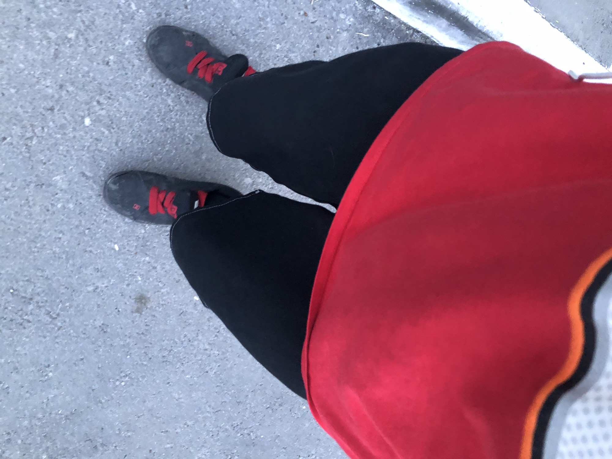 The image shows a person's legs and feet from a top-down perspective. The person is wearing black pants and dark gray athletic shoes with bright red laces. The shoes appear worn. The person is standing on a gray, gravel-like surface, possibly a sidewalk or path. In the foreground, a large portion of a red garment, possibly a shirt or jacket, is visible, obscuring the lower part of the person's torso and lap. The red garment has black and orange trim along its bottom edge. The scene appears to be outdoors. There is no indication of the specific location in Saratov, Russia.