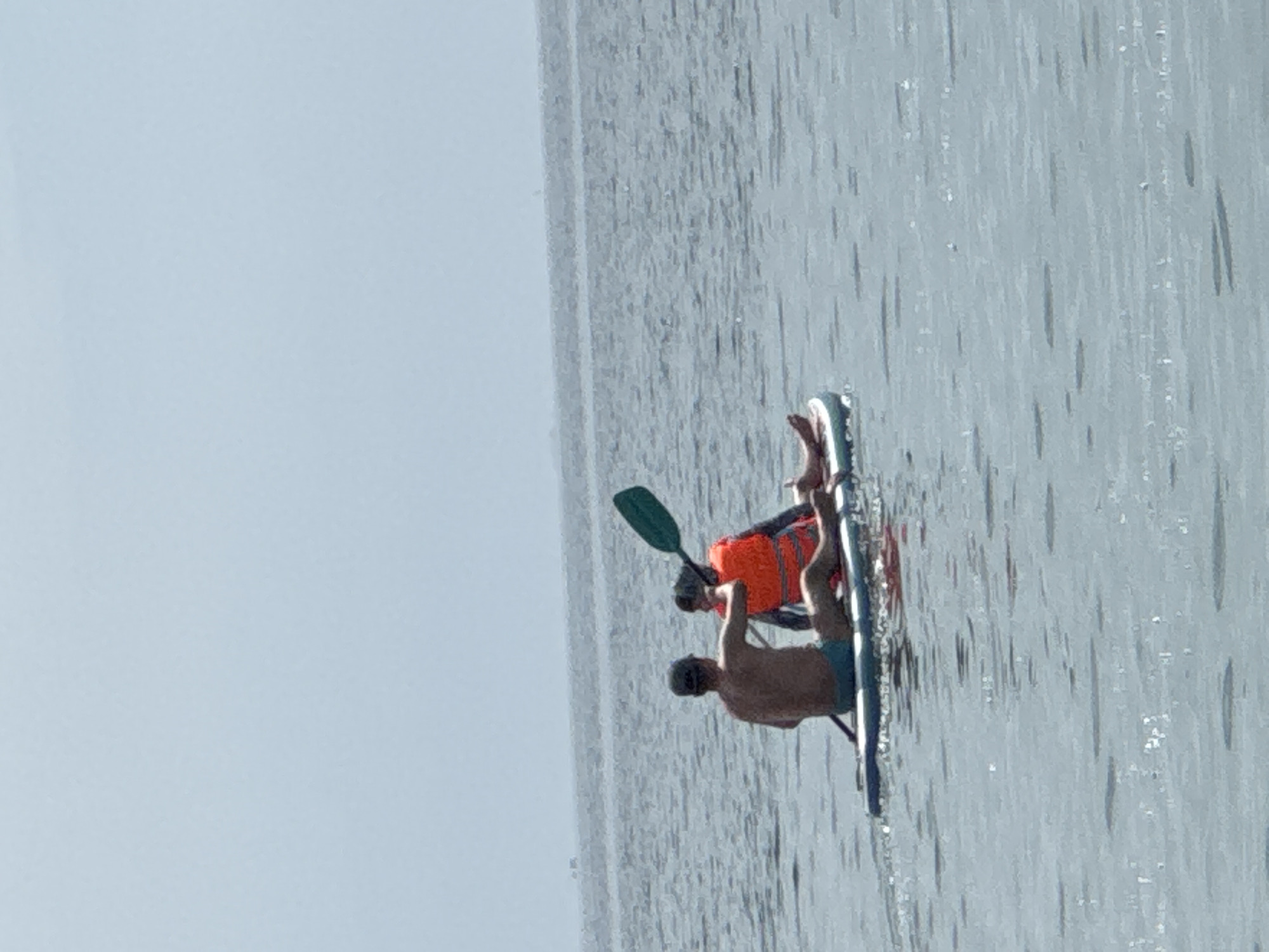 Two people are paddleboarding on the ocean in Nha Trang, Vietnam. The person in front is wearing a blue baseball cap and blue swim trunks. They are holding a paddle and appear to be steering the paddleboard. The person in the back is wearing an orange life jacket and is facing away from the camera. The paddleboard is blue and white. The water is calm and the sky is clear. In the distance, there are some buildings visible on the horizon, suggesting the proximity of the city.