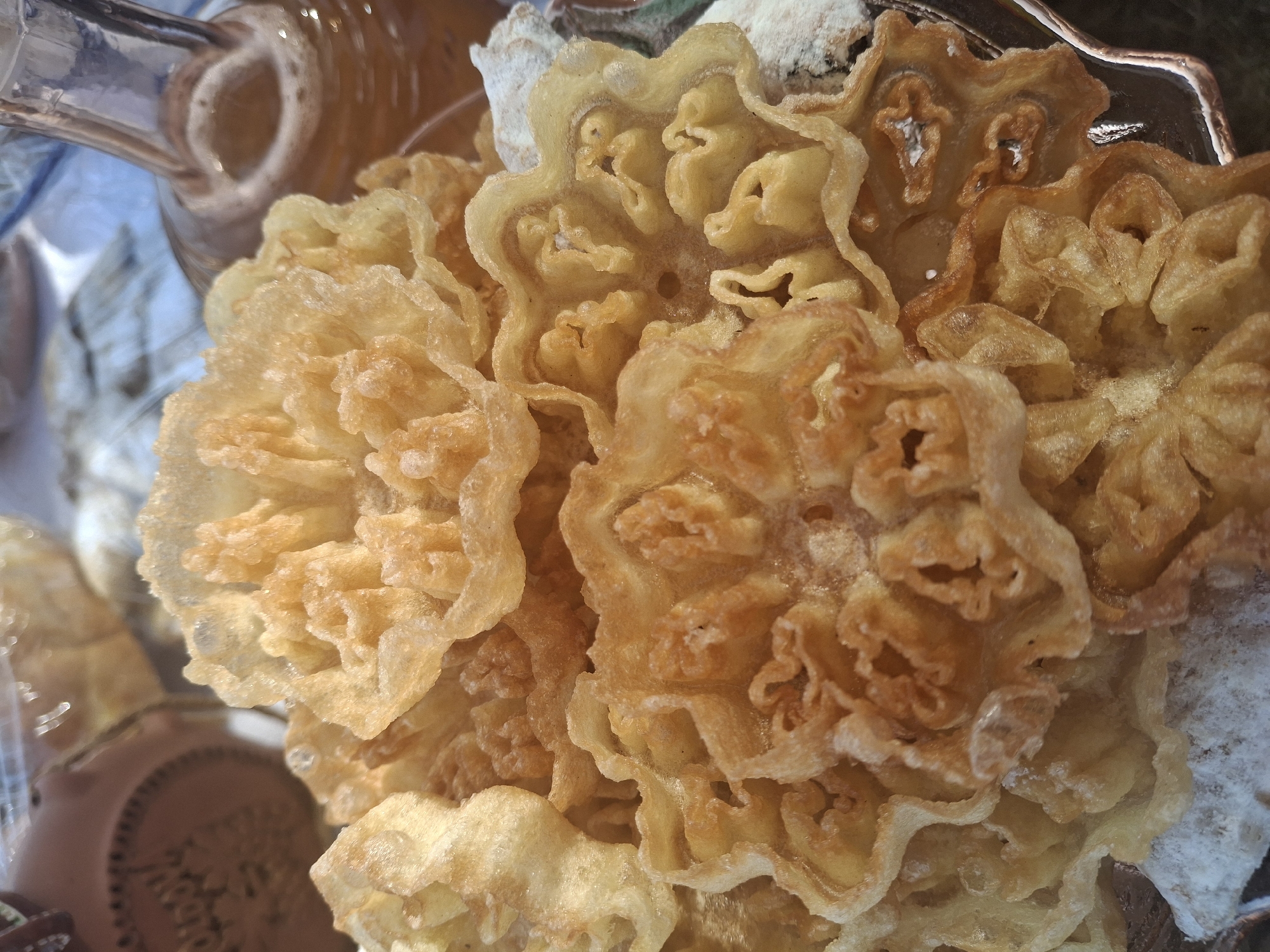 A close-up shot shows a platter of golden-brown, intricately shaped fried pastries dusted with powdered sugar. The pastries resemble delicate flowers with many ruffled petals. In the background, out of focus, a glass bottle with amber liquid and a ceramic pot are visible. The scene appears to be an indoor setting, possibly a kitchen or dining area, in Roghi, Moldova. There are no people or animals visible in the image.
