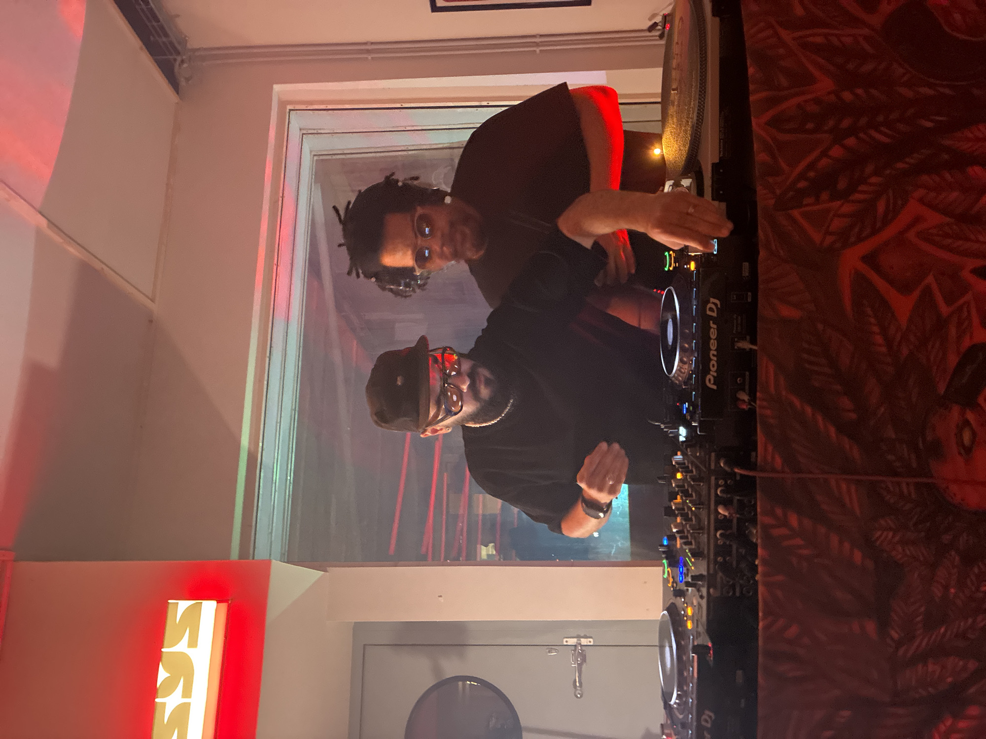 Two men are operating a DJ setup. The first man, positioned towards the front, wears a black baseball cap, glasses, a black t-shirt, and a wristwatch. The second man, standing behind, has dreadlocks, wears glasses and a black t-shirt. They are interacting with Pioneer DJ equipment in a nightclub environment, under red and somewhat dim lighting. The background features a partially obscured window. The location is Barcelona, Spain. The area to the side has a mural.