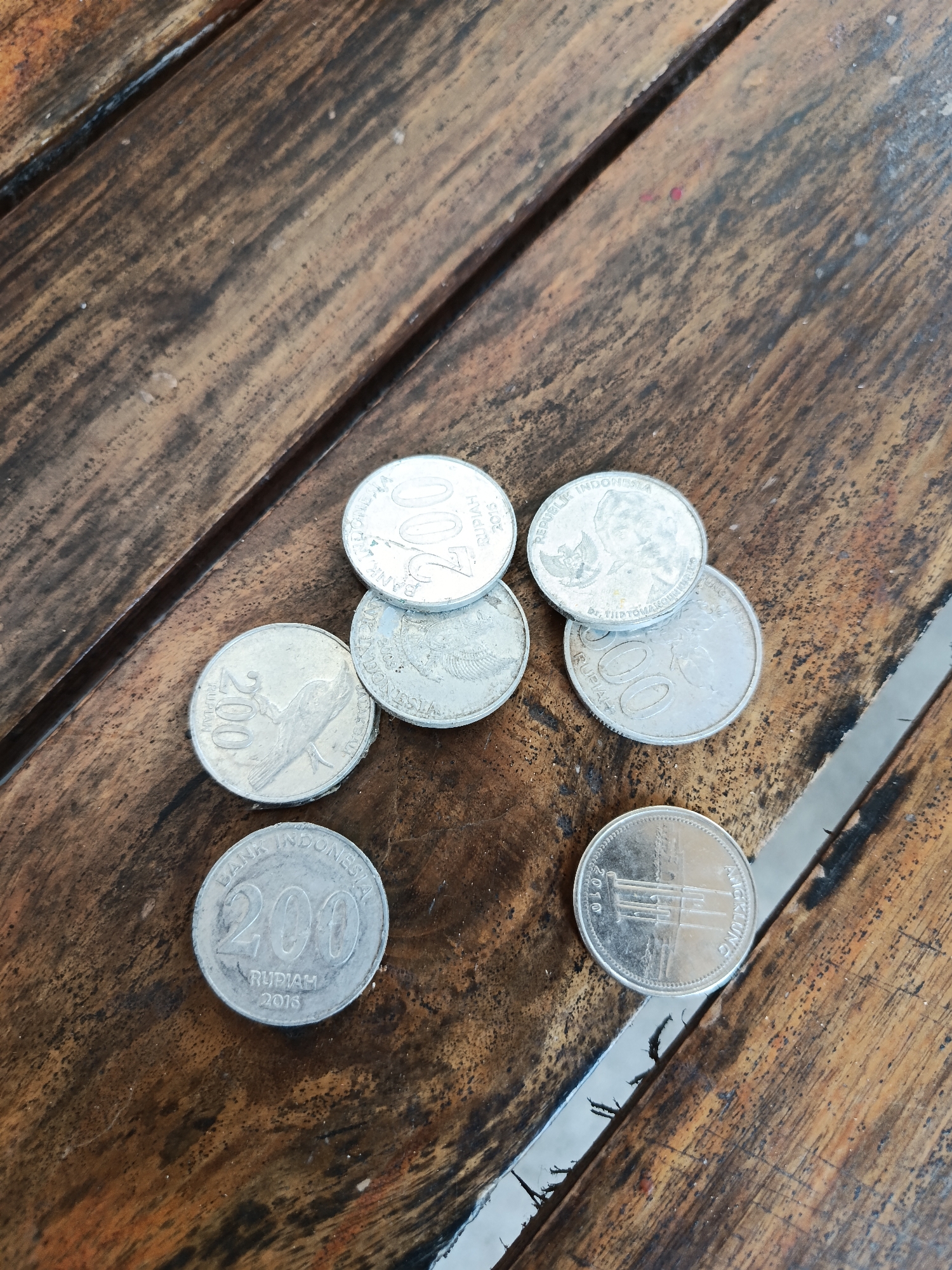 A high-angle, eye-level medium shot shows seven Indonesian coins scattered on a weathered wooden surface with prominent grain and natural imperfections. The coins are predominantly silver-toned, with some exhibiting slight tarnishing. From the top left, moving clockwise: a coin with "200 RUPIAH" and "BANK INDONESIA 2015" visible. Next, a coin displaying "REPUBLIK INDONESIA" and a Garuda Pancasila emblem, with "DR. K. R. T. RADJIMAN WEDYODININGRAT" potentially inscribed. Below that, a coin with "500 RUPIAH" and an image of a bird, possibly a kingfisher. To its right, another coin with "500 RUPIAH" and a bird, likely the same denomination. In the lower left quadrant, a coin clearly reads "200 RUPIAH" with a bird motif and "BANK INDONESIA 2013." To the right of this, a coin with the year "2010" and the word "ANGKLUNG" is visible, along with a depiction of the musical instrument. The wooden surface has a rich, dark brown color with visible cracks and dark lines suggesting natural wear and possibly wood grain. The coins are not arranged in any particular order, appearing casually placed on the table. There are no people or animals present in the scene. The lighting suggests an outdoor or well-lit indoor environment, casting subtle shadows. The location is the city of Kemiri, Indonesia.