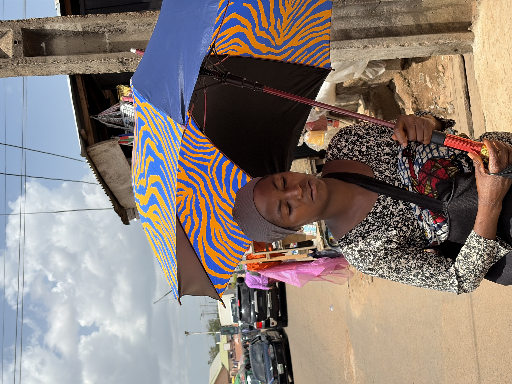 The image, taken in Jos, Nigeria, captures an outdoor street or market scene with a person holding a large, open umbrella as the central subject. The individual, likely a woman with dark skin, wears a grey headwrap and a black-and-white floral long-sleeved top over a colorful patterned fabric. Her eyes appear slightly closed, possibly squinting from sunlight.
The prominent umbrella features a distinctive pattern of alternating solid blue panels and blue panels with vibrant orange zebra-like stripes, with a black underside. The handle is reddish-brown, ending in a shiny red and gold grip.
In the background, under a bright, cloudy daytime sky, utility lines are visible. Structures with corrugated metal roofs and concrete beams suggest urban or semi-urban infrastructure. A dusty, unpaved road stretches into the distance where several dark cars are parked or moving, and blurred figures of other people can be seen. Market stalls displaying various goods, including some wrapped in pink plastic, line the roadside, indicating a bustling commercial area. The overall activity suggests daily life and commerce in a busy public space, with the umbrella likely providing shade from the sunny weather. No discernible text is visible.
