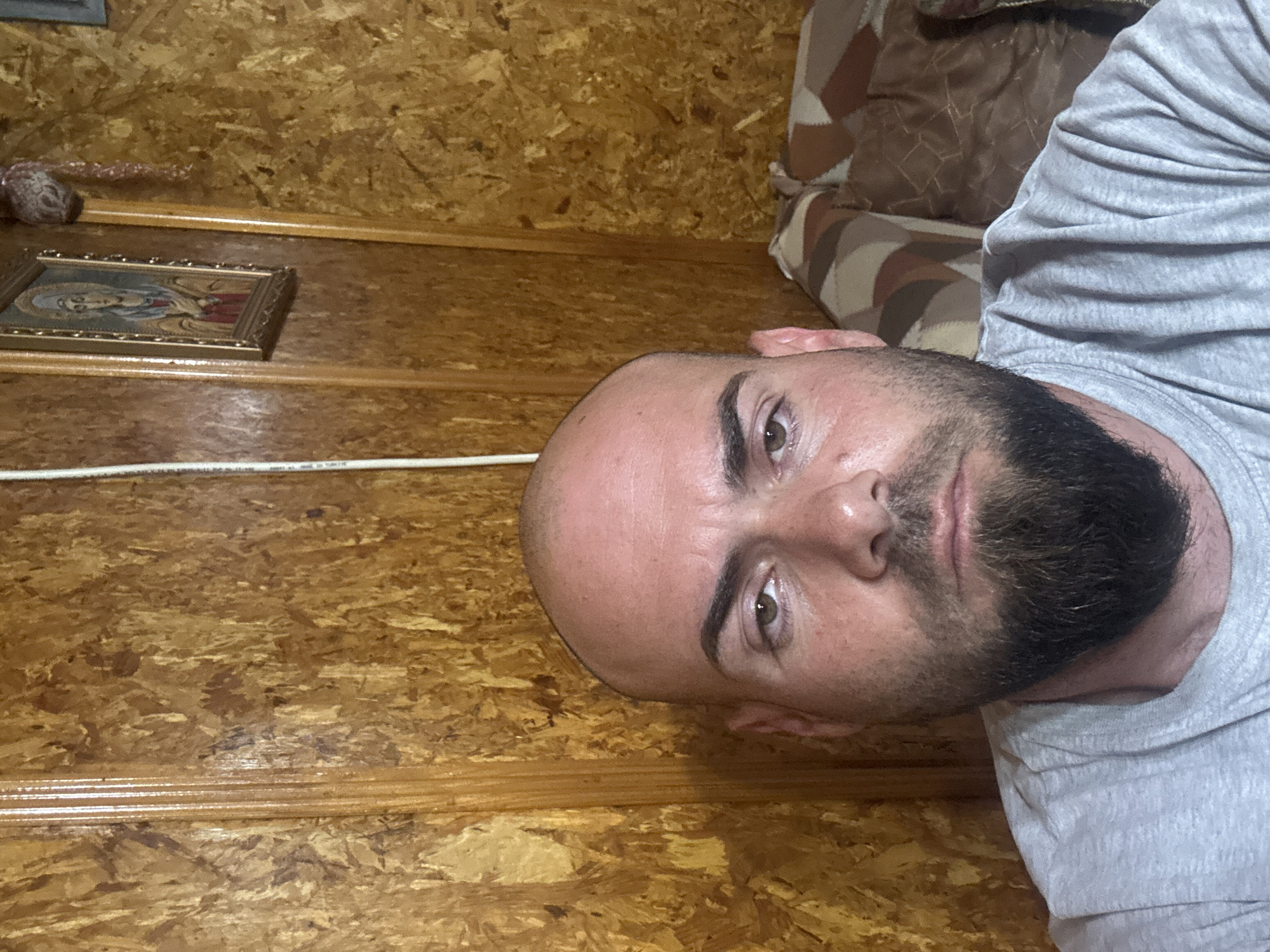 The image shows a man with a shaved head and a dark beard lying down. He is wearing a gray t-shirt. Behind him is a wall made of wood paneling. On the wall is a framed icon and an indistinct object. A white cable runs along the wall. Part of a pillow or cushion is visible in the upper right corner. The location is unknown, as there is no definitive evidence linking the image to Sarnevo, Bulgaria.