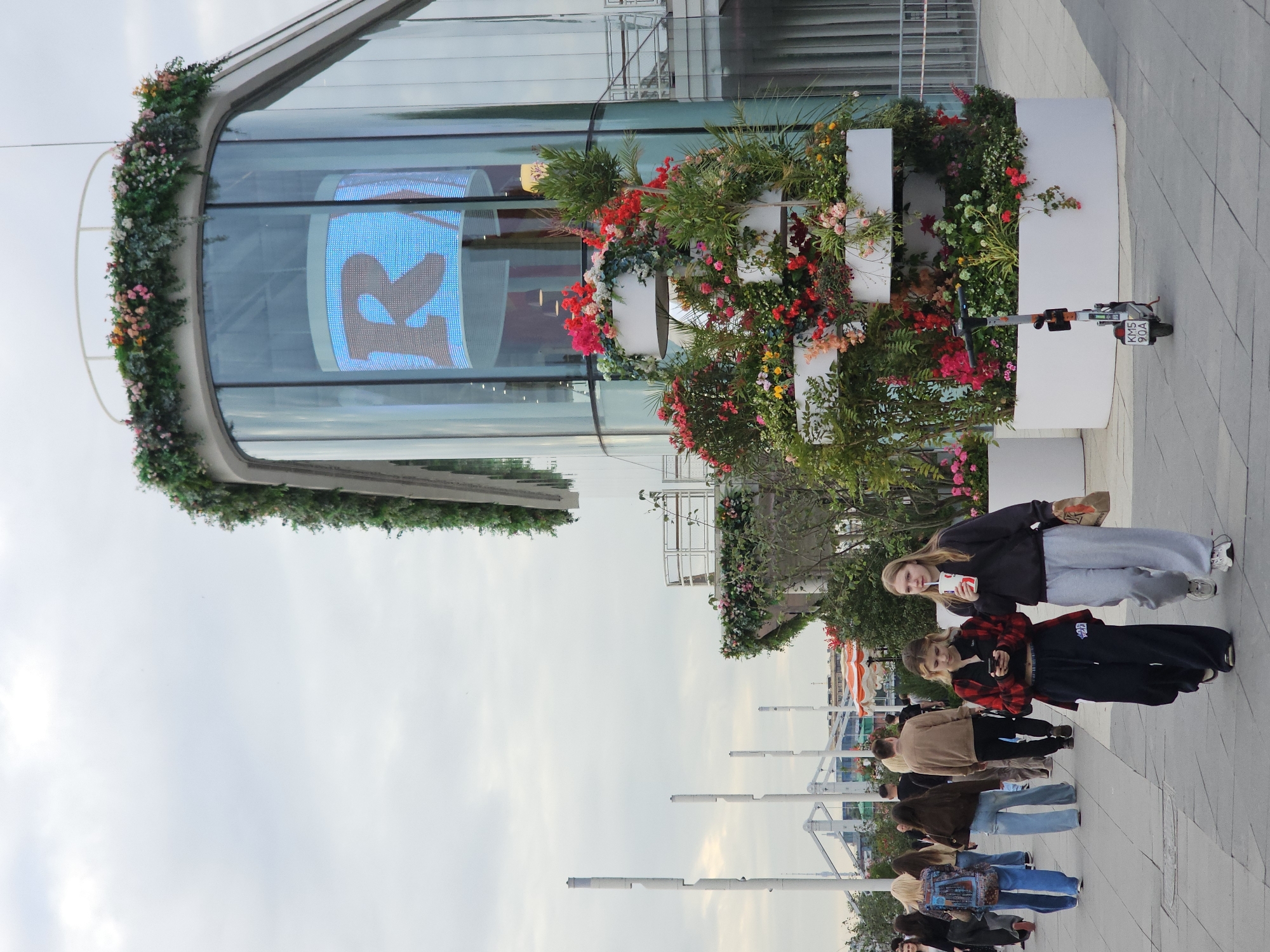 This is a photograph taken outdoors on a cloudy day. The scene depicts a group of people walking along a paved walkway in front of a modern building with large glass windows. The building is decorated with an abundance of colorful flowers and greenery arranged in planters. A white electric scooter with a license plate reading "KMS 904A" is parked near the planters.
In the foreground, several people are visible walking in a line. The two individuals closest to the camera are young women. The one on the left is holding a drink and looking towards the viewer. The woman next to her is looking down at her phone. Further back, other individuals are also walking, some carrying bags.
The building's glass windows display a large, illuminated blue and brown graphic, possibly a logo or advertisement. The overall atmosphere appears casual, and the people seem to be going about their day. The time of day is likely afternoon given the lighting.