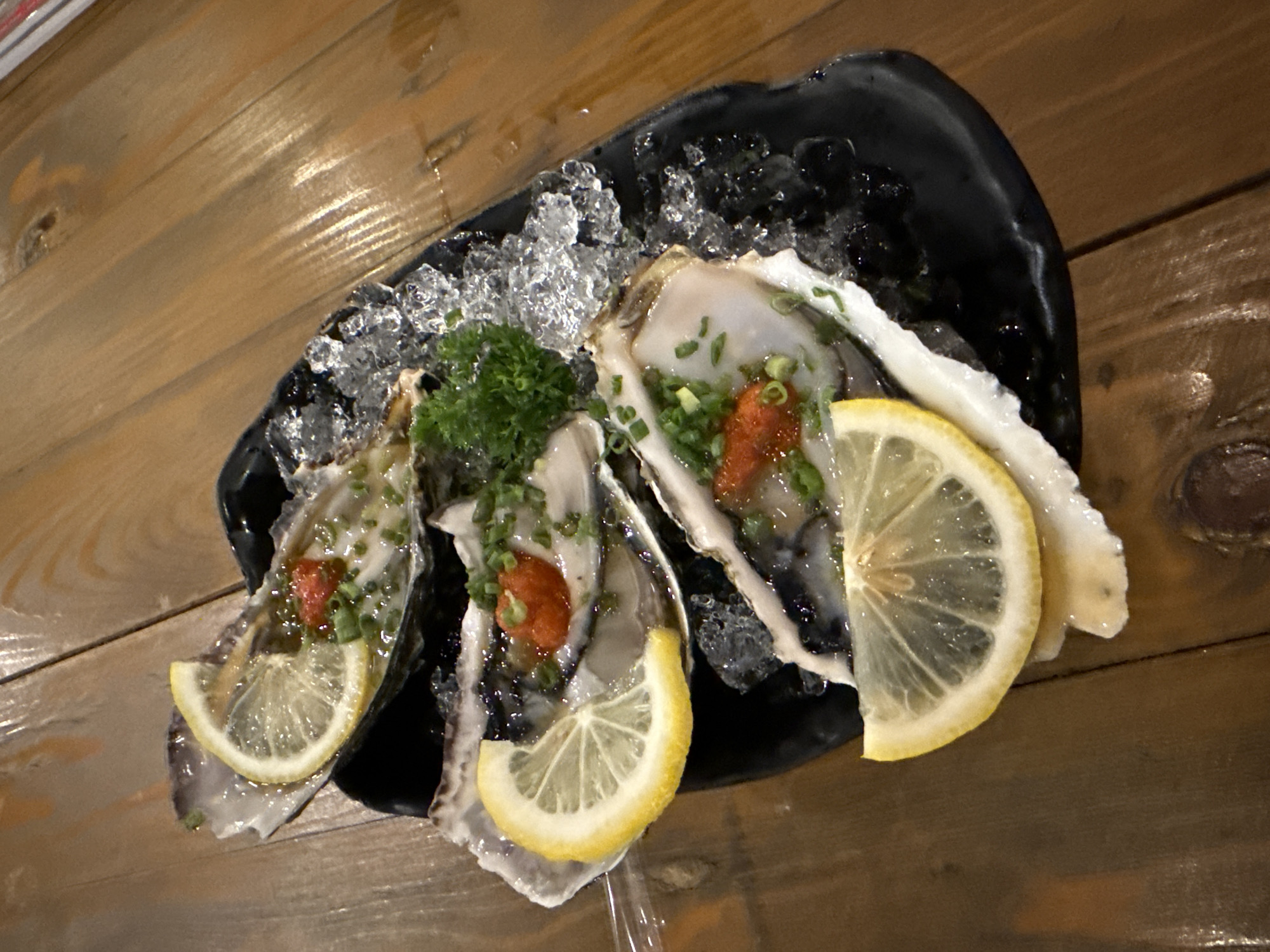 Three oysters on the half shell are presented on a dark, irregularly shaped plate filled with crushed ice. Each oyster is garnished with a slice of lemon, chopped green onions, and a reddish-orange topping. A sprig of green curly parsley separates the two oysters on the left. The plate sits on a wooden surface composed of parallel planks. The scene appears to be a restaurant setting in Bangkok, Thailand.