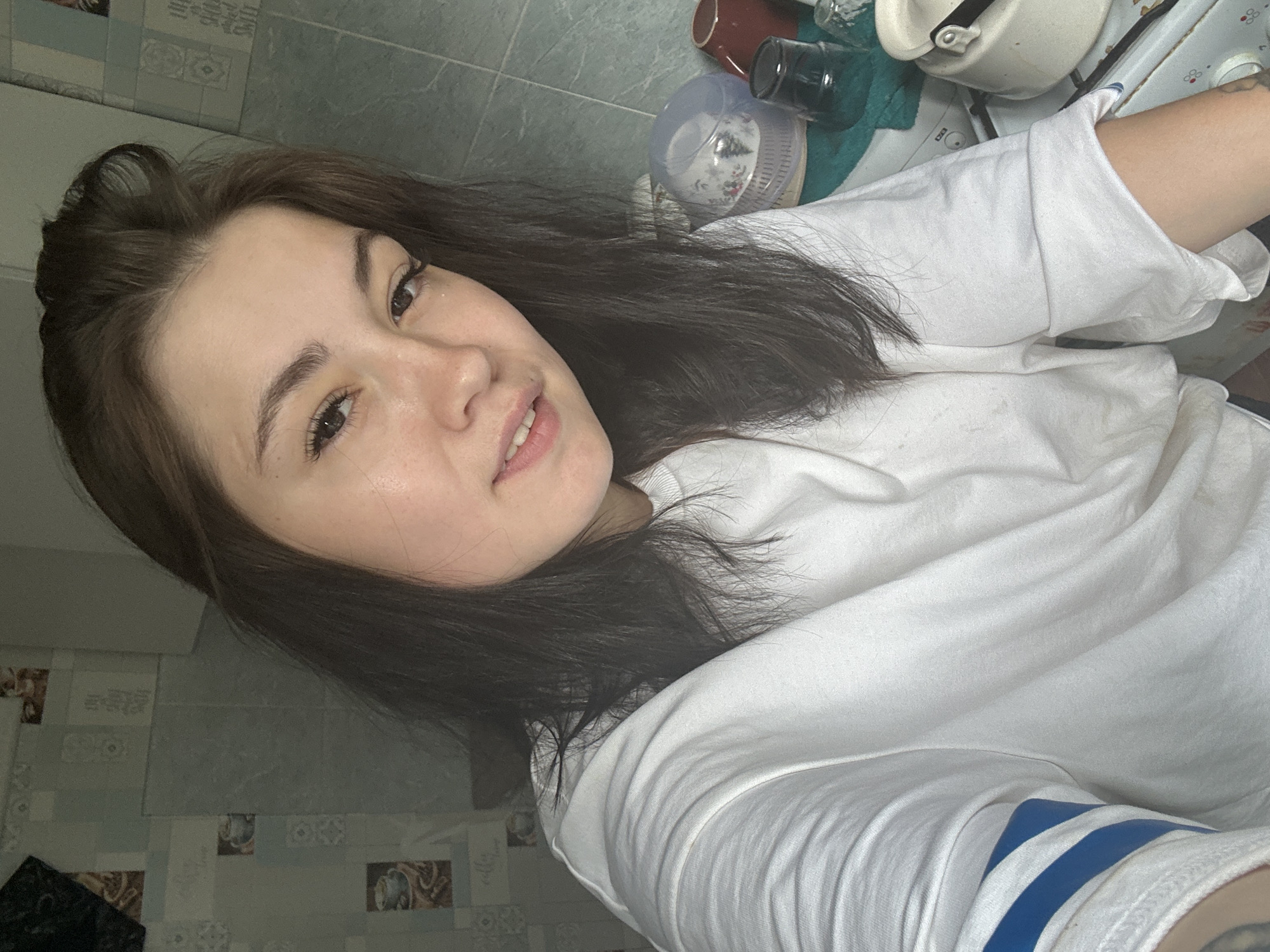 A young woman with dark hair, wearing a white sweatshirt with blue stripes on the sleeve, poses for a selfie. She is in a kitchen with blue tiled walls. On a counter to her right are several cups and a white kettle. The lighting suggests it is daytime.