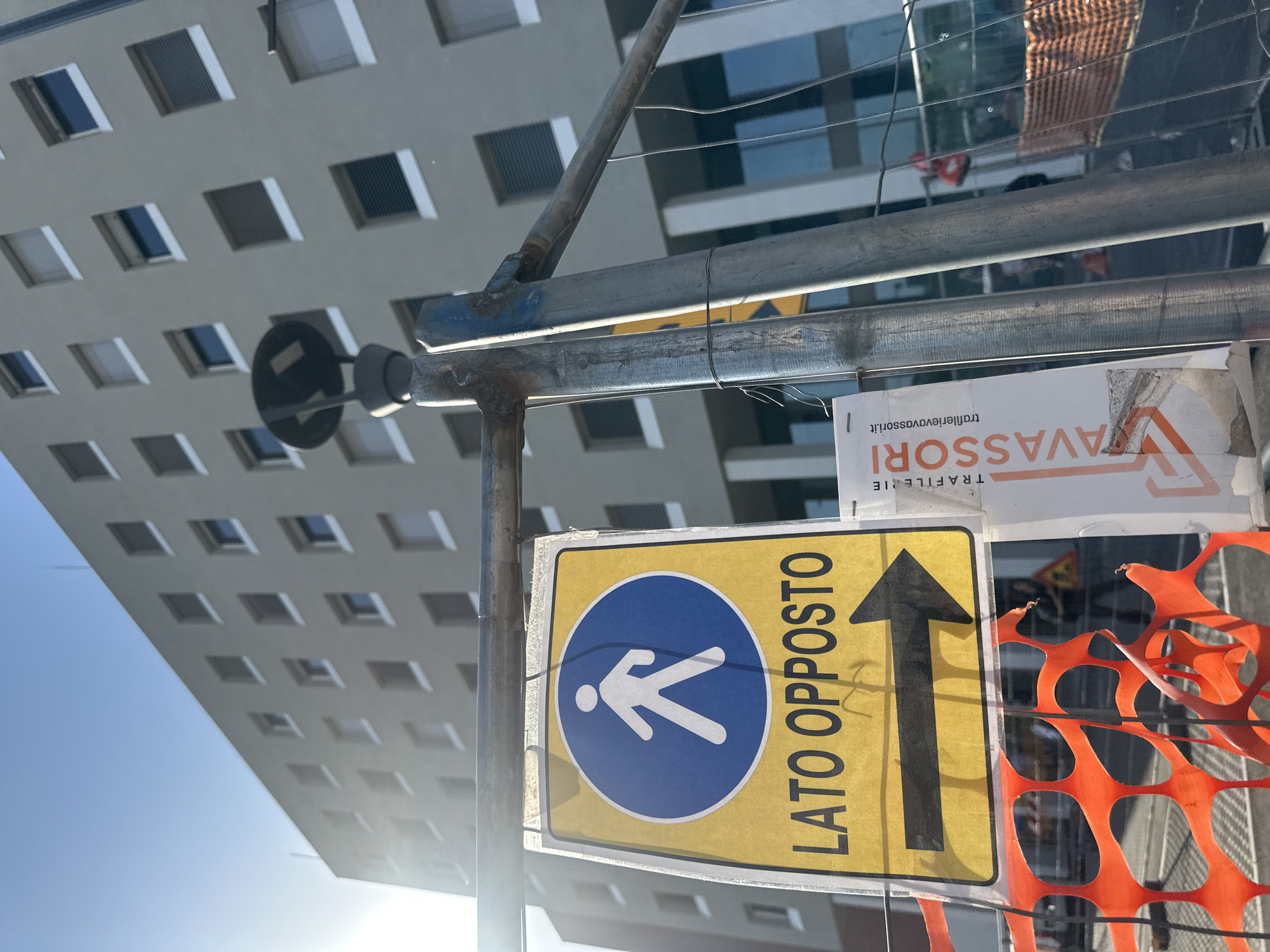The image shows a street scene in Milan, Italy. A construction site is evident, marked by an orange plastic barrier and a scaffolding structure. Affixed to the scaffolding is a yellow and blue pedestrian sign with an arrow pointing upwards, indicating the "lato opposto" (opposite side). Other signage, including a "VAVASSORI" traffic sign advertisement, is attached nearby. A tall building with a grid pattern of windows forms the backdrop, suggesting an urban setting. The sky is visible as a clear blue. No people are visible in the frame.