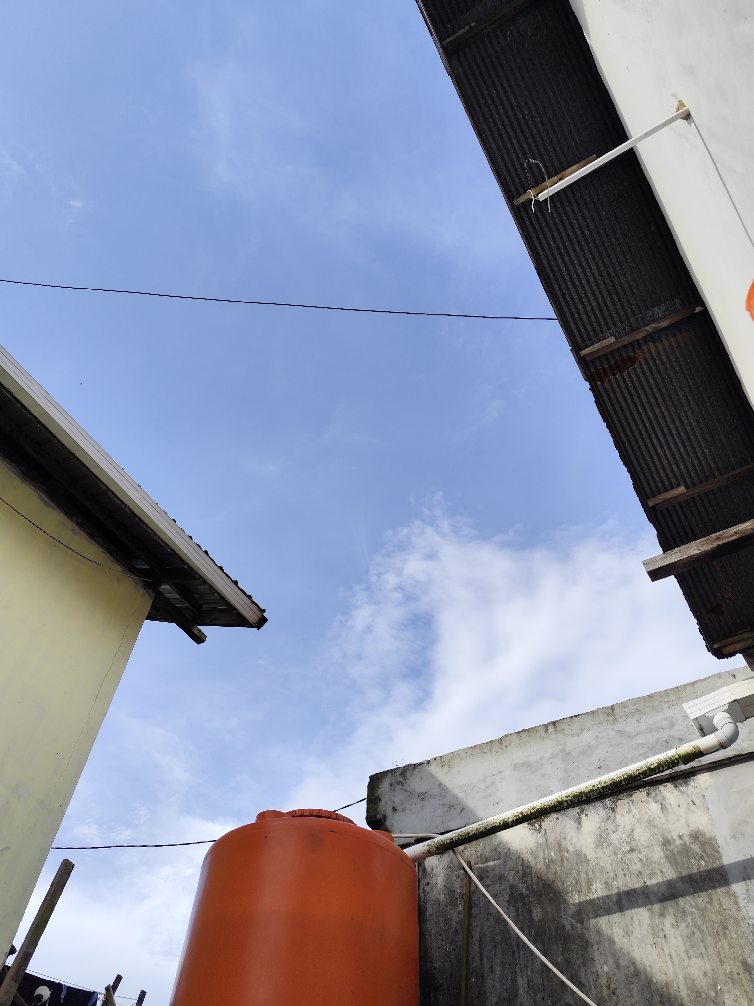 The image depicts an outdoor scene with a bright blue sky and scattered white clouds. In the foreground, an orange water tank is visible, situated next to a weathered concrete wall. A white pipe runs horizontally along the wall, showing signs of green algae growth. To the left, the pale yellow exterior of a building is partially seen, with a slanted roof made of corrugated metal. To the right, a white structure with a similar corrugated metal roof juts into the frame. A dark wire stretches horizontally across the sky in the upper portion of the image. The scene appears to be a residential or utility area, likely in a tropical climate given the clear sky and the presence of the water tank. The overall impression is one of an ordinary, functional urban or suburban environment. The specific location in Balikpapan, Indonesia, is not visually identifiable from the image itself, but the elements suggest a setting consistent with many such cities in Indonesia.