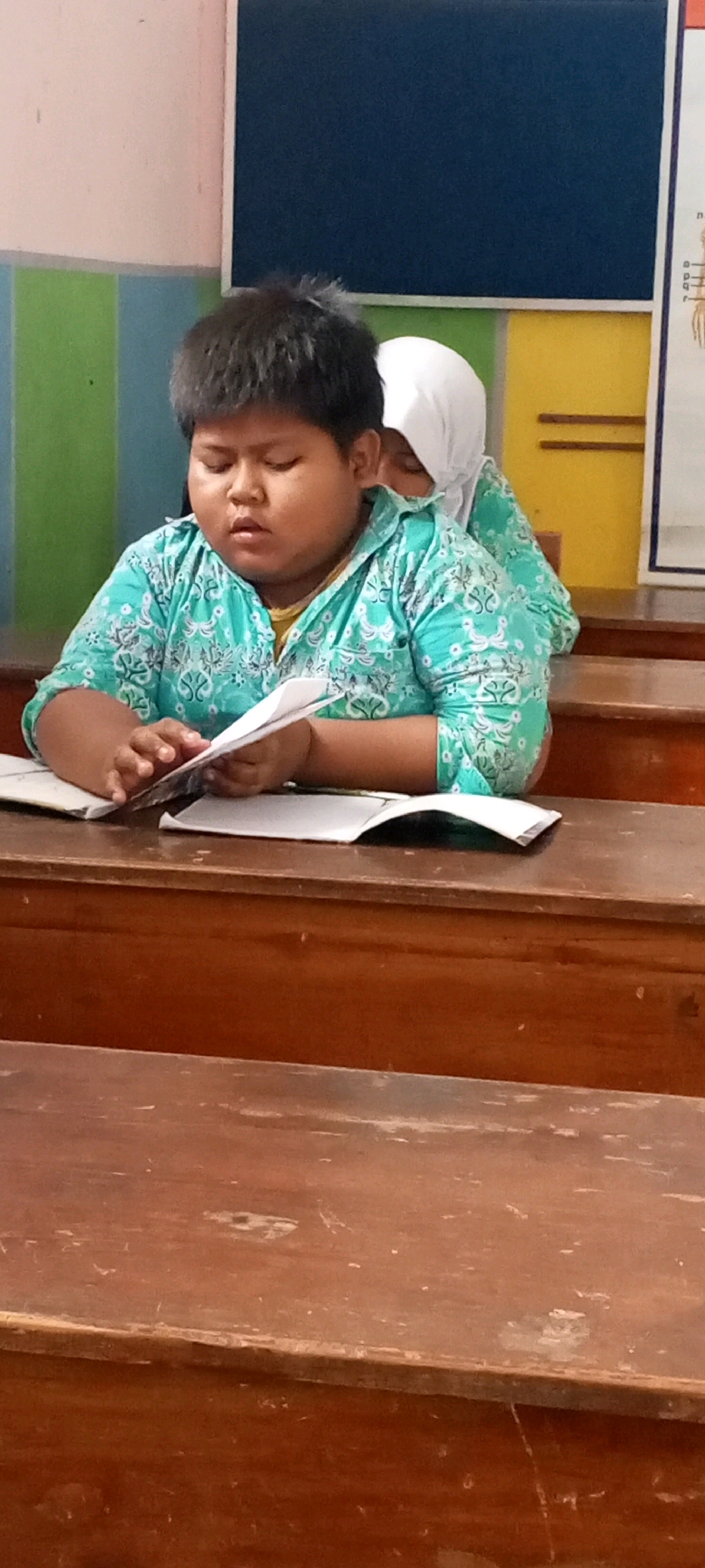 A young boy wearing a teal and white patterned shirt sits at a wooden desk in a classroom, appearing to read or study a piece of paper. His eyes are closed, and his mouth is slightly open. Behind him, partially obscured, another person wearing a white hijab is also seated at a desk. The classroom has a blue chalkboard or board in the background, with vertical stripes of pink, green, and light blue on the wall to the left. The desks appear to be old and made of polished wood. The overall scene suggests a typical classroom setting in Indonesia, likely in the city of Lumajang based on the provided context.
