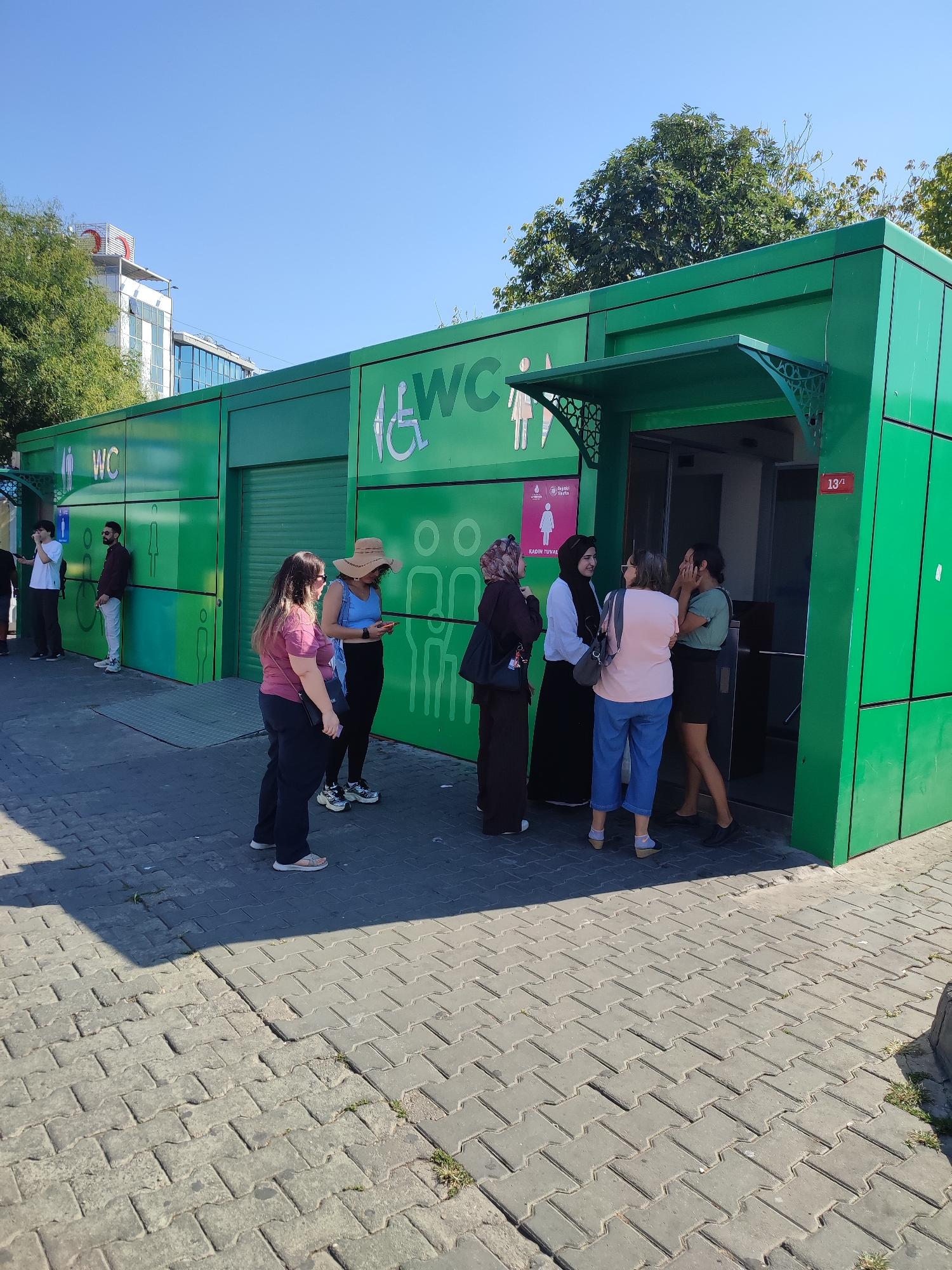 In Kadıköy, Turkey, a group of people are gathered outside a bright green public restroom facility. The building features prominent "WC" signage and symbols indicating accessibility for individuals with disabilities. Several individuals, including women wearing headscarves and a woman in a straw hat, are standing in front of or near the entrance, engaged in conversation. Another woman is leaning against the wall. On the left side of the building, two men are standing near a separate entrance, one with a backpack. The ground is paved with interlocking grey bricks, and trees and buildings are visible in the background under a clear blue sky. A sign with the number "13/" is visible on the right side of the restroom entrance.
