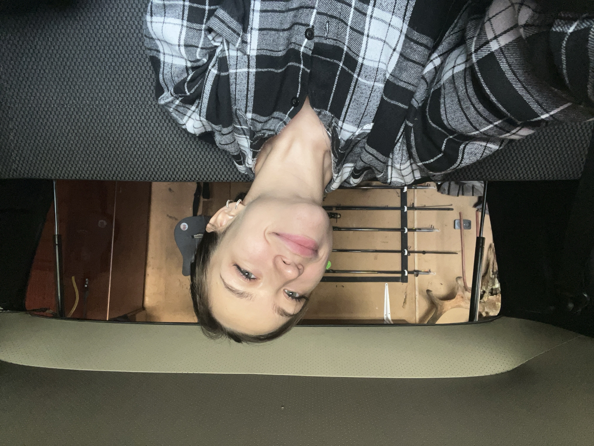 A young woman with light skin, brown hair, and blue eyes is pictured taking a selfie from inside a vehicle. She is wearing a black and white plaid shirt and is looking towards the camera with a slight smile. The background suggests the vehicle is parked in a garage or workshop, with tools and antlers visible. The interior of the vehicle, including the headliner and seats, is also visible. The image does not provide enough information to determine the location in the city of Kemul, Russia.