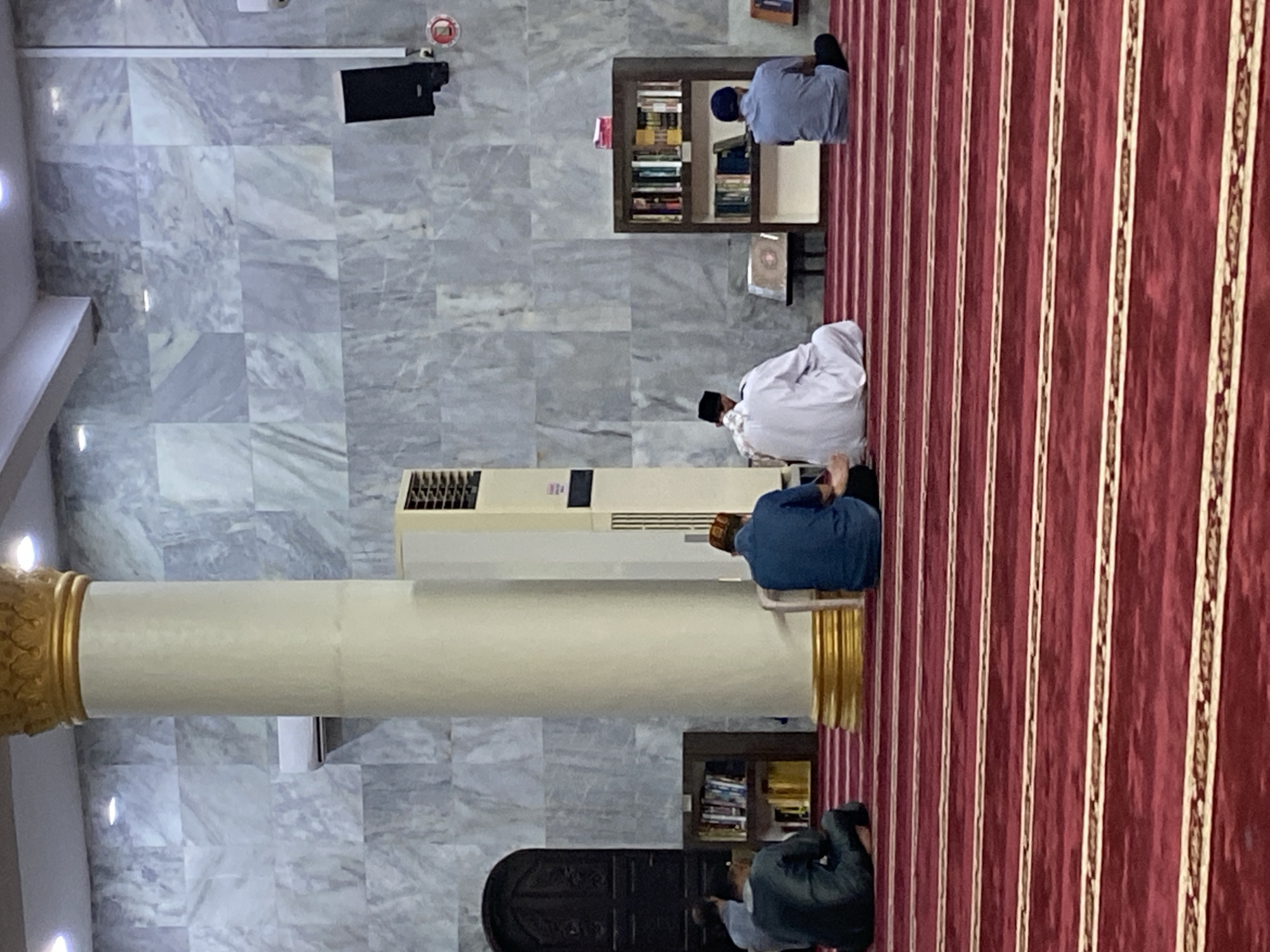 The image shows the interior of a mosque in Lhokseumawe, Indonesia. Several men are seated on a red carpet with white stripes, facing away from the camera. The walls are covered in gray marble-patterned tiles, and a large white pillar with a gold capital stands prominently in the center. A tall, cream-colored air conditioning unit is visible to the right of the pillar, and a dark wooden door is on the left. Bookshelves filled with books are placed against the wall to the left and right of the center. A black speaker is mounted on the wall in the upper right corner.