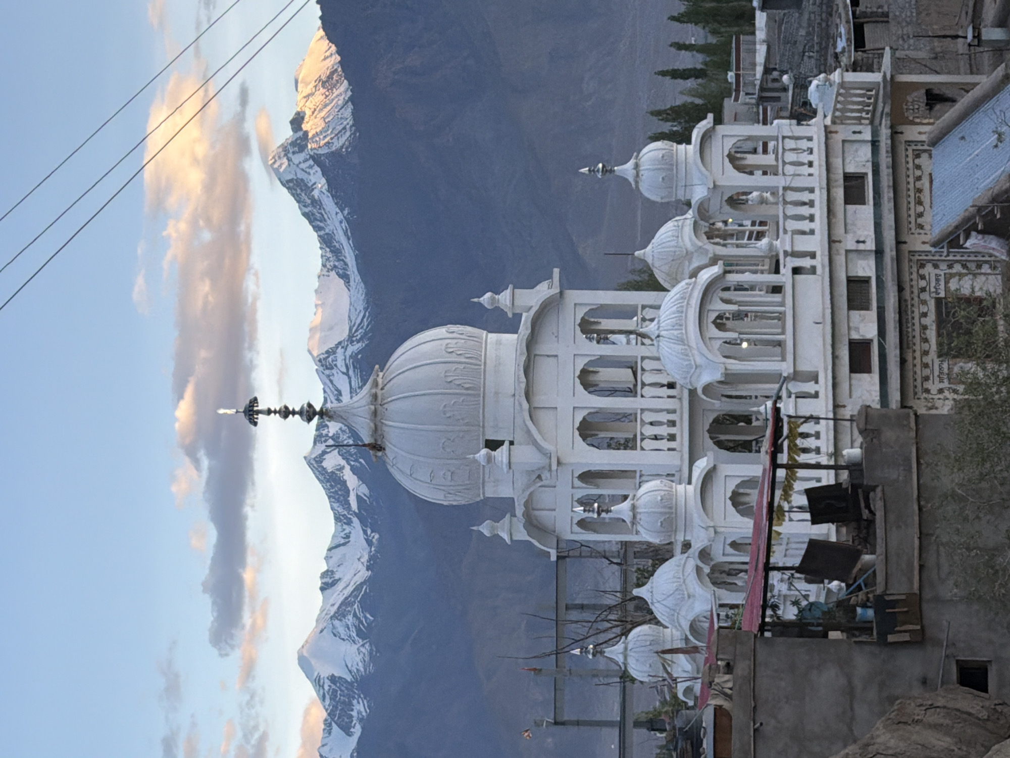 The image shows a white, multi-domed building, likely a Gurdwara or a Buddhist monastery, in the city of Leh, India. The building has intricate architectural details, including arches, minarets, and decorative carvings. It is situated in a mountainous region, with snow-capped peaks visible in the background. The sky is partly cloudy, with a gradient of blue and pink hues, suggesting either sunrise or sunset. In the foreground, there are several other buildings, some appearing to be older and made of stone, with a few trees and utility poles visible. There are no people or animals clearly discernible in the image. The overall scene is tranquil and picturesque, capturing a blend of architectural beauty and natural grandeur.