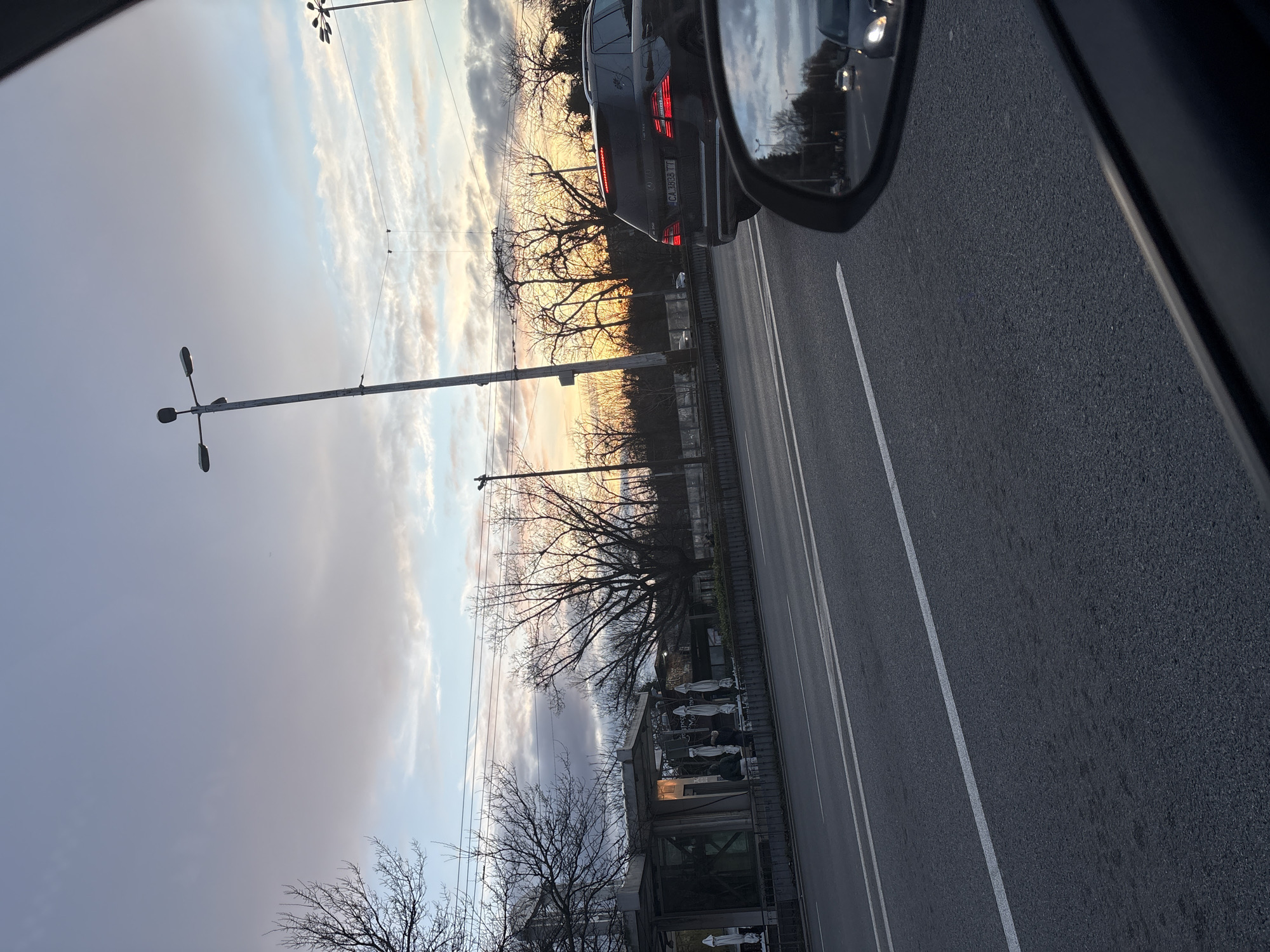 The image shows a view from a vehicle, likely a car, traveling on a road. A side mirror is visible in the top right corner, reflecting other vehicles. To the left of the road is what appears to be a cemetery with visible gravestones and possibly small shrines. The sky above features a mix of clouds and some clear areas, potentially indicating a sunrise or sunset. Utility poles line the road. The road itself is paved and has lane markings. The scene suggests an urban environment with a cemetery adjacent to a road. Based on the visible license plate "CA 1828 TT", the location is in Sofia, Bulgaria.