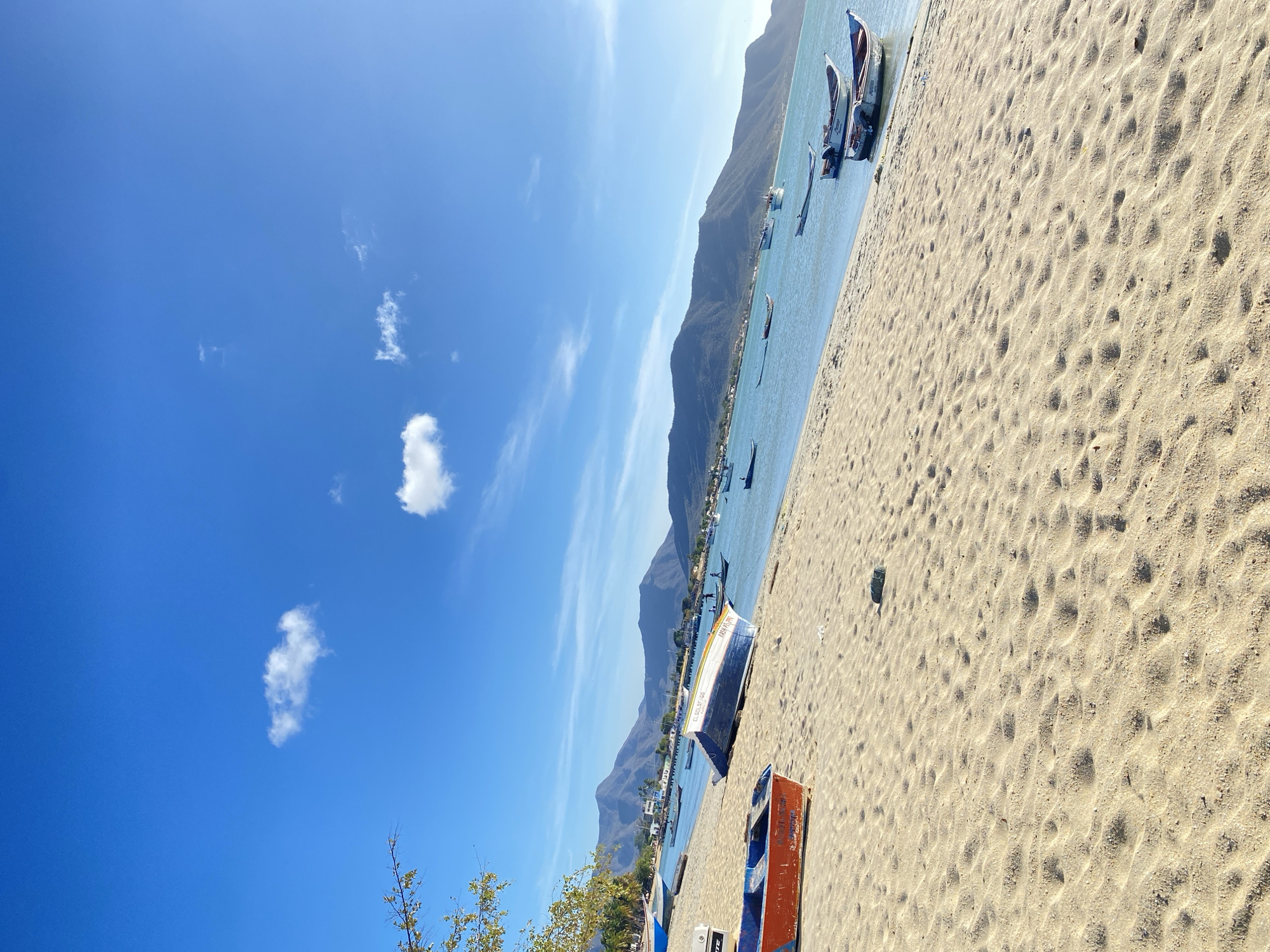 This image depicts a coastal scene in Juan Griego, Venezuela, under a clear, bright blue sky with two prominent white cumulus clouds. The foreground consists of a sandy beach, characterized by ripples and visible footprints. Several small, wooden fishing boats (peñeros) are present on the beach and in the water.
In the lower-left, an orange and blue fishing boat, partially visible, rests on the sand, showing the name "VIANNA". To its right, a blue and white fishing boat, labeled "EL GOLFO", is overturned on the sand. Farther left, another blue boat is partially visible on the sand. A small, dark object, possibly debris, is located on the sand in the lower-right quadrant.
The midground features calm, light blue-green water of a bay or lagoon, extending towards the horizon. Multiple similar fishing boats are moored or floating in the water, some close to the shore, others further out. In the mid-right, two blue and white boats float near the water's edge, one identified as "Princesa" and "CARLA".
The background is dominated by a range of dark, undulating mountains stretching across the horizon. A small settlement with visible buildings and sparse vegetation is situated along the shoreline at the base of these mountains. No individuals are visible, and no specific actions or interactions are depicted within the frame. The scene is brightly illuminated, indicating daylight hours.