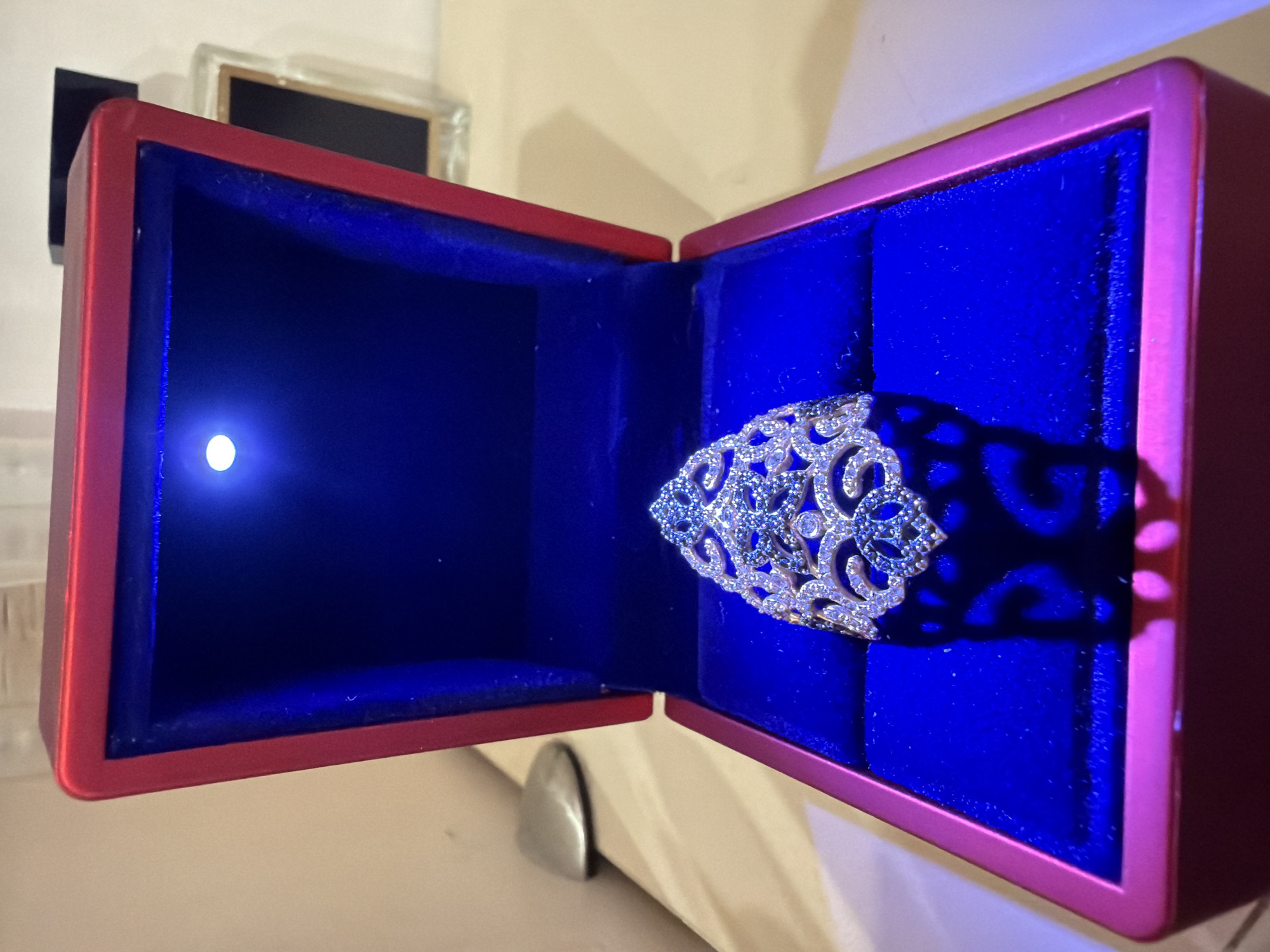 A close-up, eye-level shot captures a ornate ring placed inside a red, velvet-lined jewelry box. The box is open, revealing the ring centrally positioned on the plush blue interior. A small, bright LED light illuminates the ring from the top of the box. The ring itself features intricate metalwork with a symmetrical, swirling design adorned with numerous small, clear and blue gemstones. The background is softly blurred, showing a light-colored surface with what appears to be a perfume bottle in the upper right corner and a metallic object on the left. The overall lighting suggests an indoor setting, possibly a dressing table or a display area. The image does not provide any specific details or context related to the city of Kyiv, Ukraine.