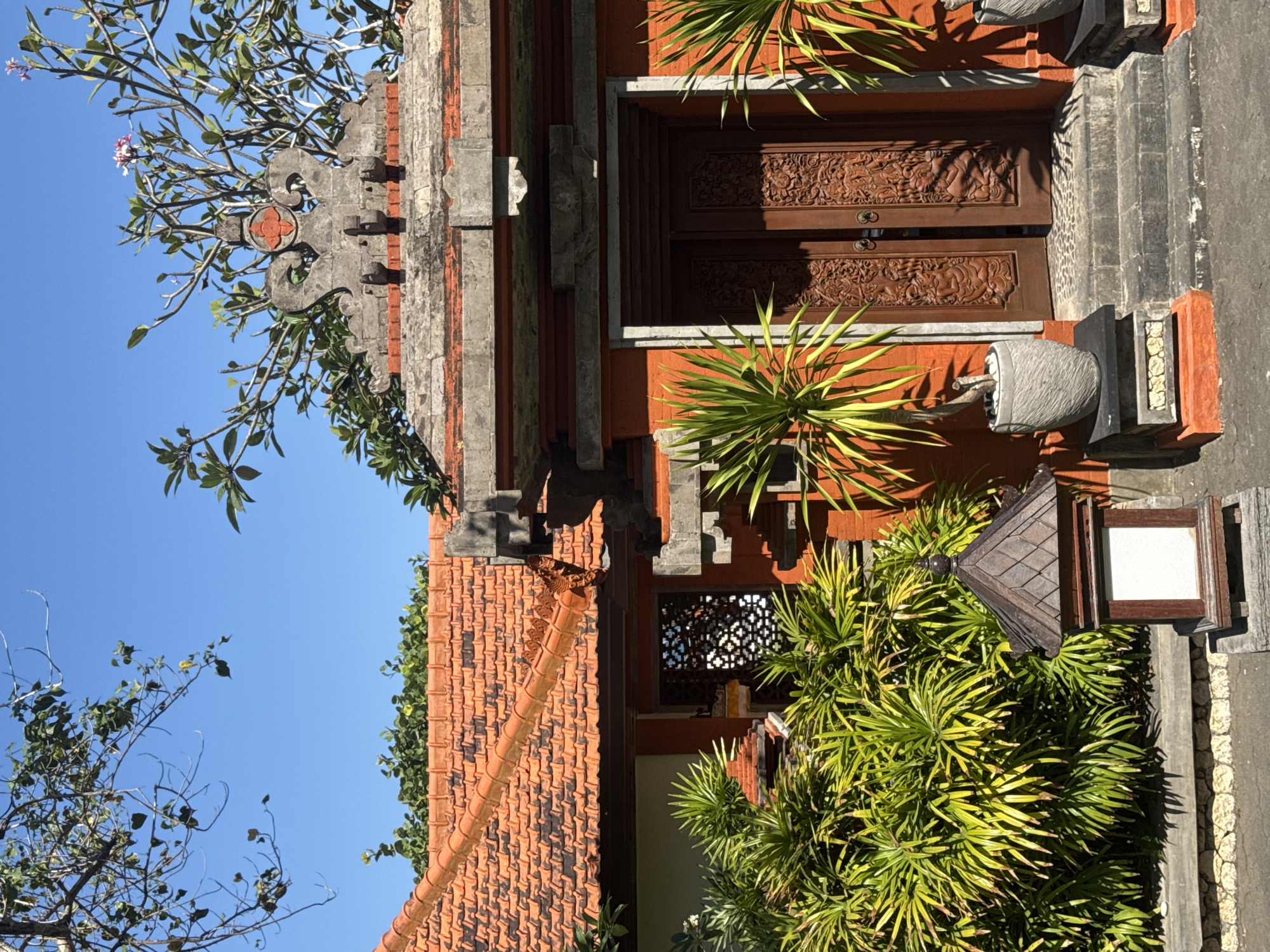 The image shows the entrance to a traditional Balinese building in Kuta Selatan, Indonesia. The building features an orange tiled roof, a red-painted wall, and intricately carved wooden double doors. Flanking the entrance are two potted plants with long, green leaves. Above the doors, a decorative stone carving sits atop the wall, with branches of a flowering tree reaching into the clear blue sky. A small, dark wooden lantern with a white panel sits to the left of the entrance. The ground in front of the entrance is paved with stone.