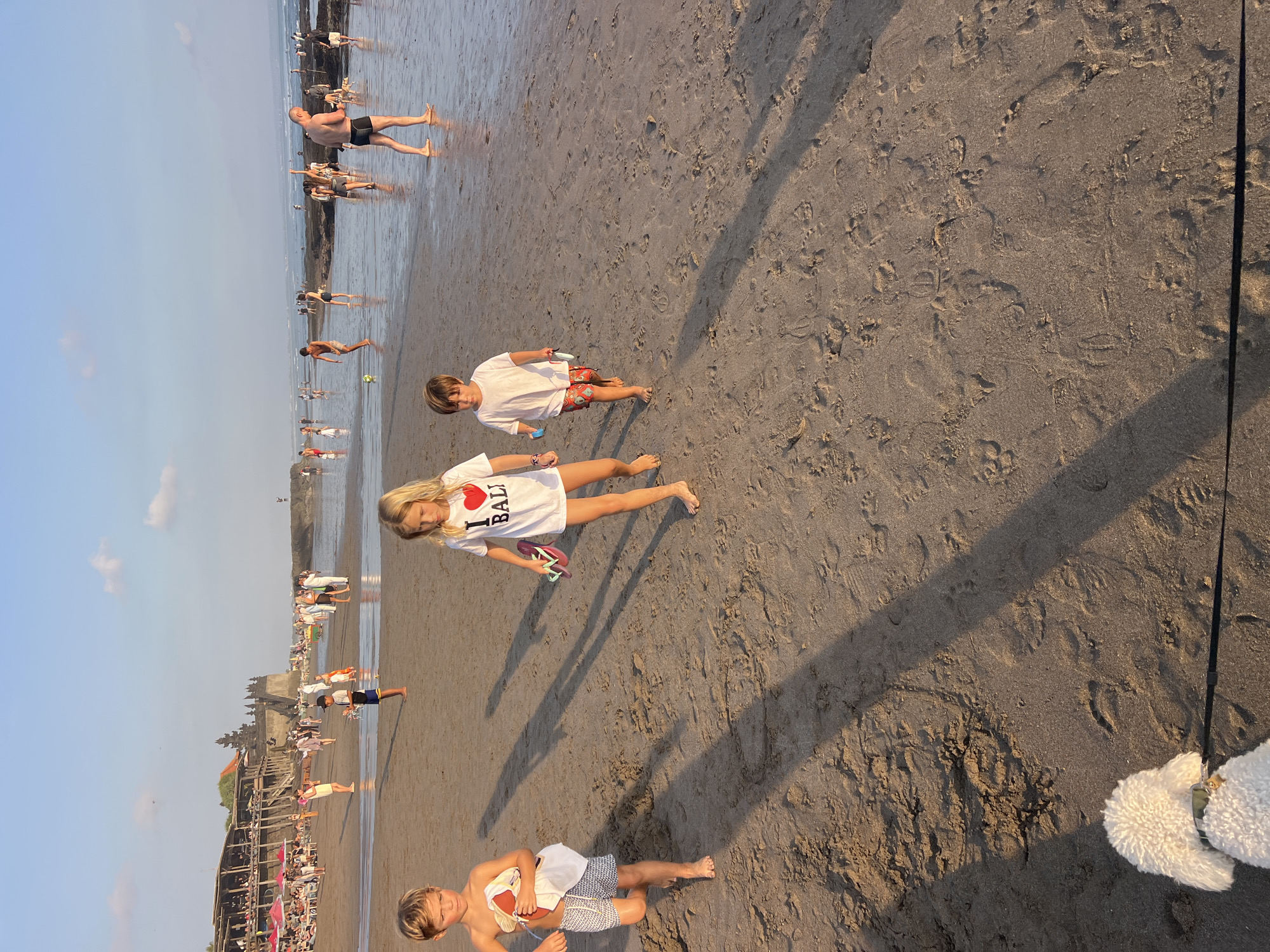 The image shows a beach scene in North Kuta, Indonesia, during what appears to be late afternoon or early evening. The sky is a pale blue with a few wispy clouds, and the setting sun casts long shadows across the sand.
In the foreground, three children are walking on the wet sand near the water's edge. A young boy on the left, wearing patterned shorts and holding a white cloth, walks towards the viewer. To his right, a girl with blonde hair, wearing a white t-shirt that says "I LOVE BALI" and holding pink flip-flops, walks in the same direction. Further to the right, another boy in red patterned shorts and a white t-shirt is also walking towards the viewer. A white poodle is in the bottom left corner, on a leash held by an unseen person.
In the background, many other people are visible on the beach and in the shallow water. Some are standing, some are walking, and some appear to be playing. Further in the distance, there are buildings and structures, including what looks like a temple complex and a restaurant or bar with thatched roofs, situated on a rocky outcrop. The ocean is visible in the far background. The sand is a light brown color and shows many footprints.