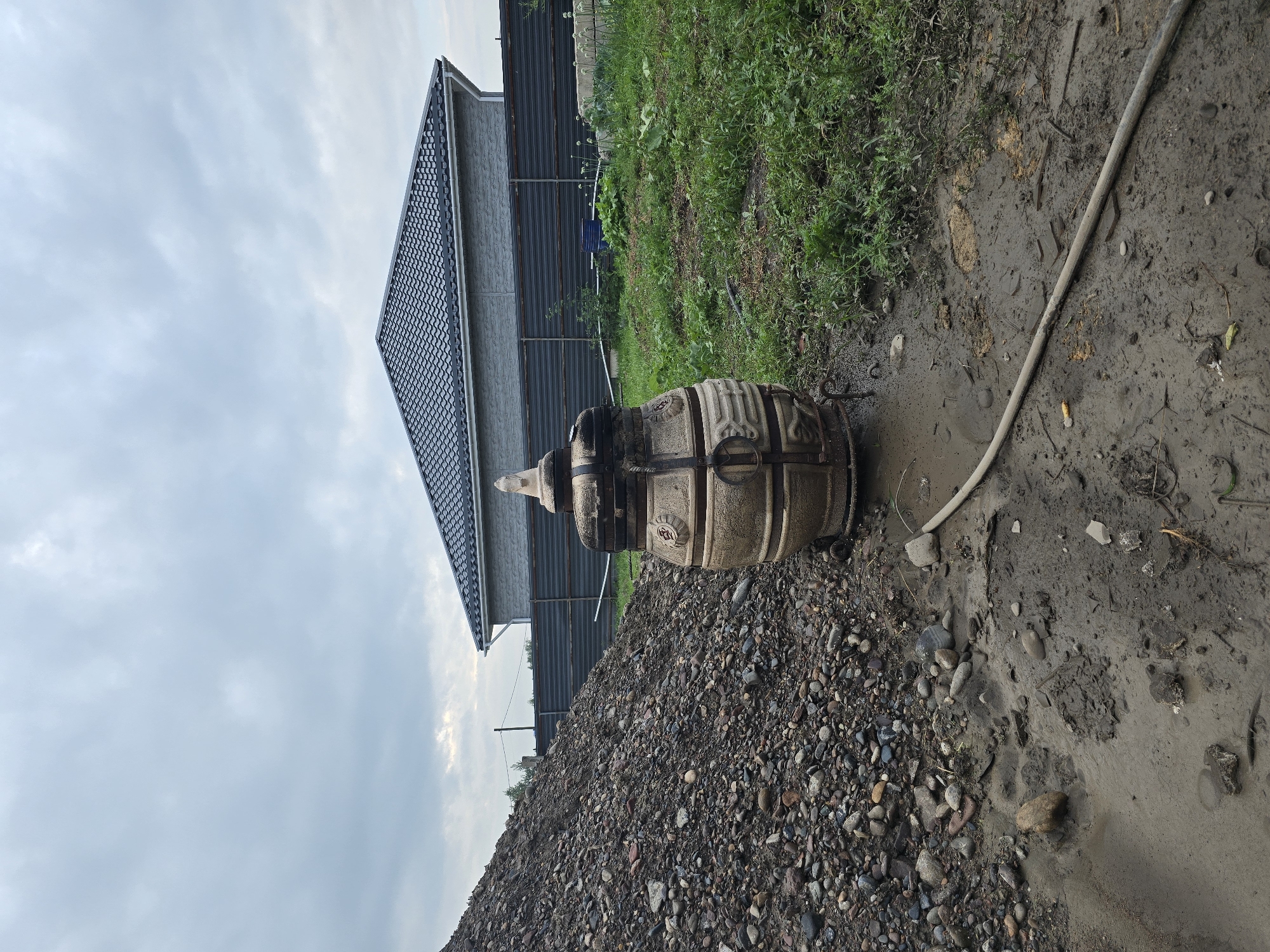The image depicts an outdoor scene in Ермолаево, Russia, on a cloudy day. In the foreground, a pile of gravel is on the left, and a tandoor oven stands in the center. The oven is made of beige and dark metal, with intricate decorative elements. It is placed on a metal stand and appears to be used or recently used, with some soot marks visible on its upper section. The ground around the oven is muddy and uneven, with a hose lying across it. To the right of the oven is a patch of green grass and vegetation. In the background, a grey house with a dark tiled roof is visible, along with a dark metal fence. Power lines and a utility pole are also present in the distance. The overall impression is one of a construction or renovation site, or an area undergoing landscaping. There are no people visible in the image.