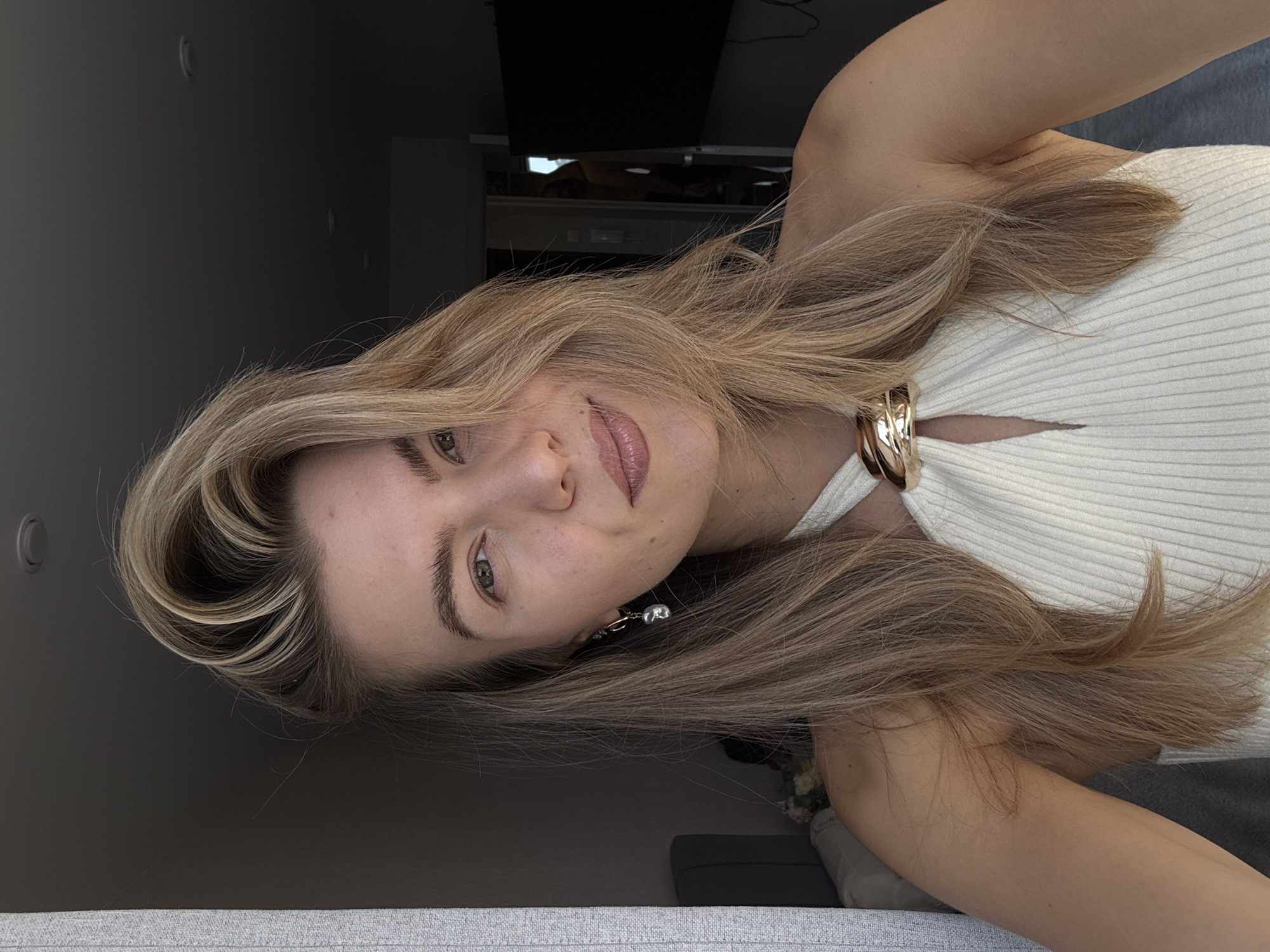 A woman is centered in a selfie-style portrait with her head tilted to the right. She is wearing a white ribbed top that has a gold detail at the neckline. Her hair is long, light brown, and styled with a wave falling across her forehead. She is wearing a small earring with a pearl accent on her left ear. The background shows a muted gray wall, a television, and furniture, suggesting an indoor setting. The location is the city of Unknown, Unknown.
