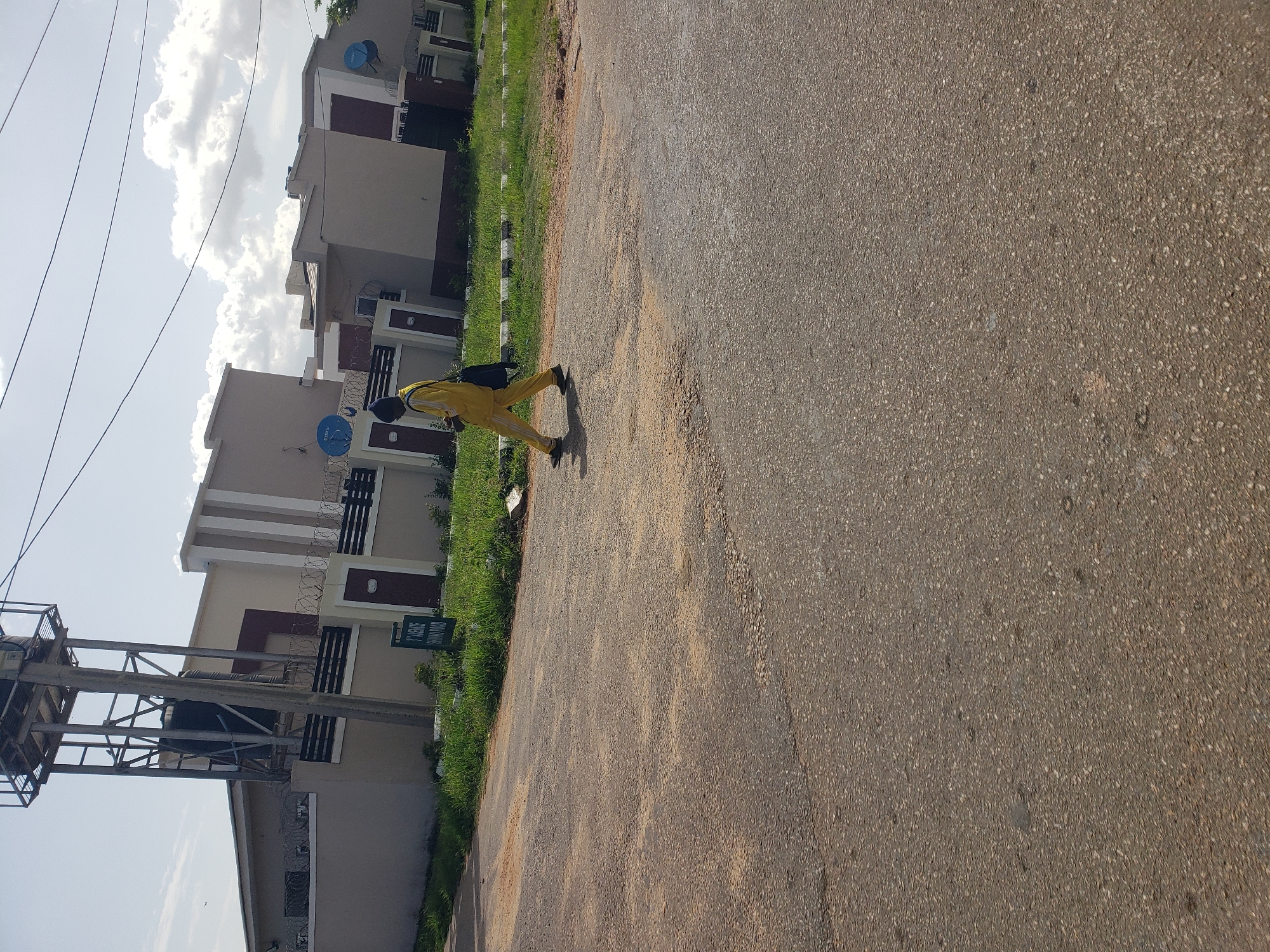 The image depicts a street scene in Gura Top, Nigeria. A person wearing a yellow tracksuit and a blue beanie walks across the paved road, carrying a dark bag over their shoulder. The road appears to be made of asphalt and shows signs of wear. In the background, a row of modern residential houses with beige and white facades are visible. Some of the houses are equipped with satellite dishes. A large water tower stands to the left of the houses. A sign indicating "3rd AVENUE SHIMA ROAD" is planted in front of the houses. The sky is partly cloudy, and vegetation, including grass, lines the side of the road and the property boundaries. The scene appears to be set during daylight hours, with the sun casting shadows.