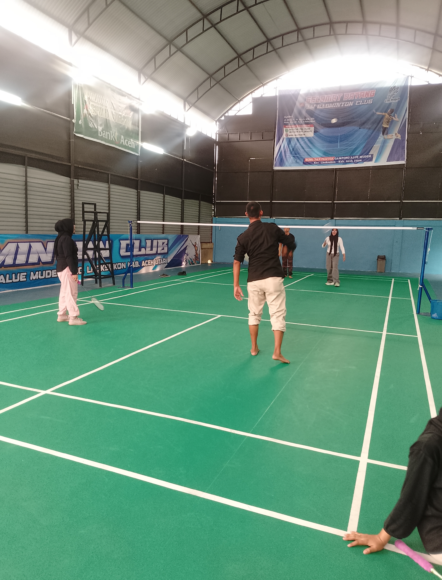 The image depicts an indoor badminton court with four individuals playing a game. The court is green with white boundary lines. A badminton net is stretched across the center of the court.
In the foreground, on the left side, a person wearing a black headscarf and pink pants is positioned, holding a badminton racket. On the right side, another person in a black shirt and light-colored shorts is in motion, facing away from the camera. In the background, two other individuals are visible on the court, one in a white headscarf and grey pants, and another further behind in a brown shirt.
Banners are visible on the back wall of the court. One banner is green and displays the text "Bank Aceh". Another larger blue banner announces "SELAMAT DATANG DI AM BADMINTON CLUB". This banner also includes text in smaller font, mentioning "ALAMAT: Jl. Tgk. Ibrahin B. Aceh Km. 102, Alue Mudem Kec. Lhoksukon", and "BUMI DAN PEMUDA GAMPONG ALUE MUDEM, Kec. Lhoksukon - Kab. Aceh Utara". This information confirms the location as Alue Mudem village, within the Lhoksukon sub-district, North Aceh Regency, Indonesia.
The environment is an indoor sports facility with a corrugated metal roof and industrial lighting. There are also metal structures and equipment associated with the badminton court, such as a referee's stand and net poles.
