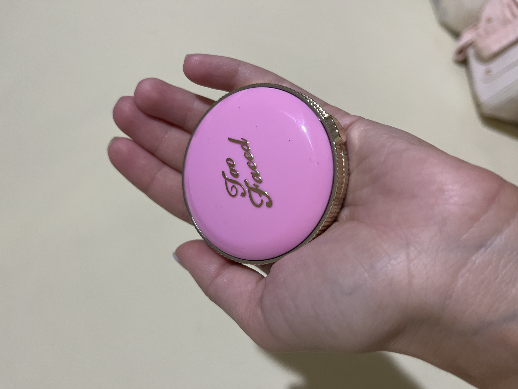An image shows a person's hand holding a pink compact with gold trim. The compact has the "Too Faced" logo in gold script. The background is a plain, light-colored surface. The location is Unknown, Unknown.