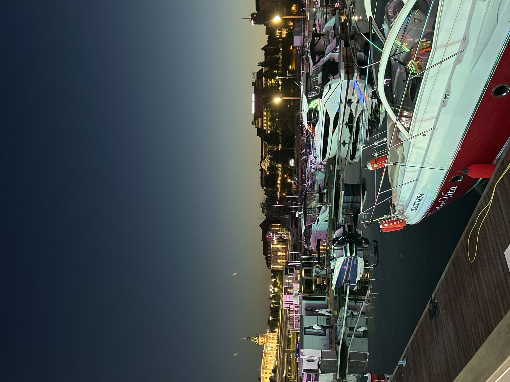 This is a photograph of the marina in Szczecin, Poland, taken at dusk. The sky is a deep gradient of dark blue and lighter blue, with a hint of yellow at the horizon. The foreground is dominated by a wooden pier. Moored to the pier are several boats, some of which are lit with colorful LED lights. One prominent white and red boat in the foreground has "POLO01K5X" and "Dulce Vita" written on its side. In the middle ground, there are several floating structures that appear to be modular cabins or houseboats. A few people are visible on the pier and on one of the floating structures. In the background, the cityscape of Szczecin is illuminated. Buildings with ornate architecture and glowing windows are visible, along with streetlights and a prominent tower structure. The water in the marina reflects the lights from the boats and the city, creating a shimmering effect.