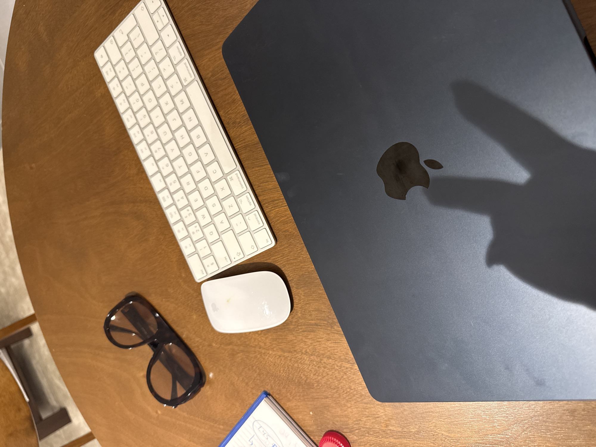 This is a top-down shot of a desk with a laptop, keyboard, mouse, sunglasses, notebook, and other small objects. The desk appears to be made of wood. The laptop is a dark color with the Apple logo visible. A hand is casting a shadow on the laptop. The keyboard is white and compact. The mouse is white and oval-shaped. The sunglasses are dark-rimmed with a brown tint. A blue notebook and a red object are at the bottom of the image. The overall scene is a workspace arrangement on a wooden desk. There is no information in the picture to conclude that it is in Dubai, United Arab Emirates.