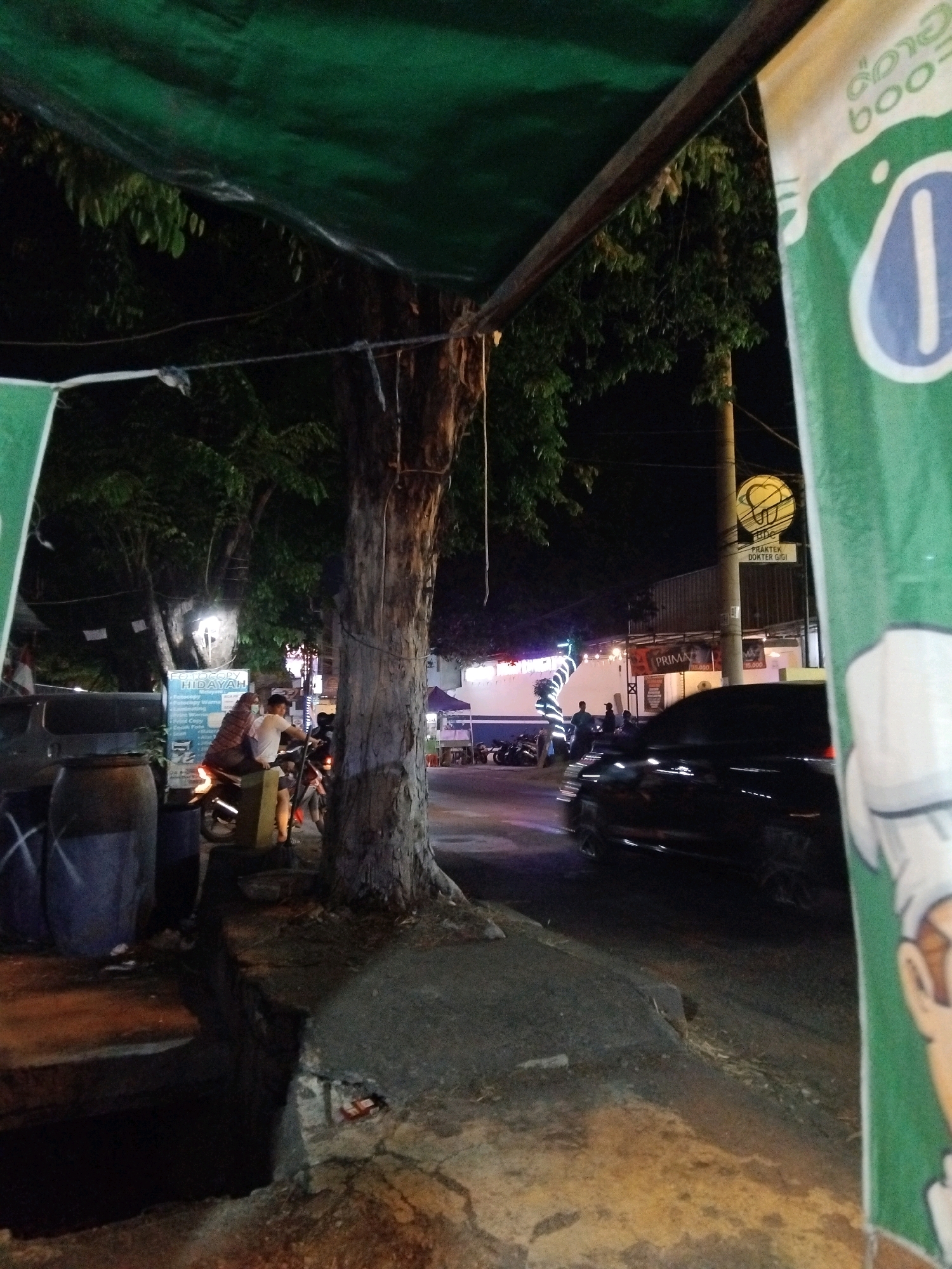 The image shows a street scene at night in Wedoro, Indonesia. A large green tarp is stretched overhead, supported by a wooden beam. To the right, a portion of a tall, green banner with white text and graphics is visible, depicting what appears to be a cartoon character's head and shoulders wearing a chef's hat, and possibly some beverage branding.
In the foreground, to the left, are two large, dark, cylindrical objects, possibly barrels or water tanks, with white crosses marked on their sides. A concrete curb separates the sidewalk from the road. A large, rough-barked tree trunk is prominent in the center of the image, rising vertically.
On the sidewalk, a person is sitting on a motorcycle, facing away from the camera. They are wearing a white t-shirt and dark shorts. Another person is partially visible, also on a motorcycle, with their back to the camera. A sign that reads "FOTOCOPY & HIDYAH" with additional text below is attached to a pole behind the people on the motorcycles.
The road is visible in the background, with a black car driving on it. Streetlights illuminate the scene, casting shadows and creating a dimly lit atmosphere. Further down the street, buildings and shops can be seen, some with illuminated signage. A yellow circular sign with a tooth symbol and text is mounted on a pole to the right. The overall impression is a busy street corner at night, with commercial activity and traffic.