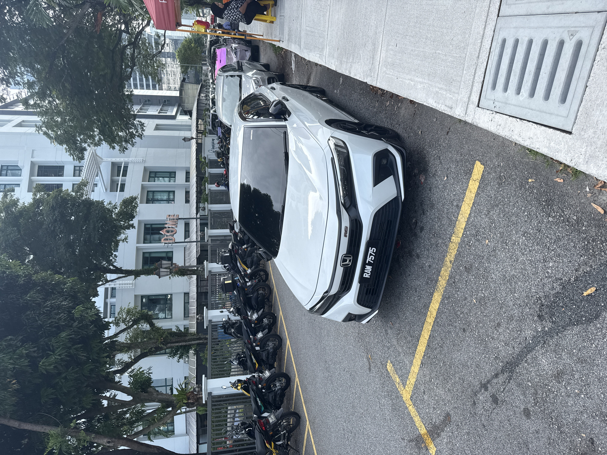 This image captures an outdoor urban scene in Kuala Lumpur, Malaysia, depicting a street or parking area bordered by buildings and a sidewalk.
In the foreground and midground, a white Honda City RS car is prominently featured, parked diagonally on asphalt. Its license plate, "RAM 7575", is clearly visible on the front bumper. The car is sleek and modern, with black accents on its grille and wheels. To the left of the car, a row of motorcycles is parked, many of them scooter-style, behind a metal fence or railing. Yellow lines mark the parking spaces on the asphalt.
To the right, a concrete sidewalk runs parallel to the street, featuring a large, rectangular grey storm drain cover with vertical slats. Further along the sidewalk in the background, a few people are visible, some sitting on what appears to be a yellow outdoor bench or at a table, suggesting an outdoor cafe or waiting area. Other cars are parked further down the street on the right.
The background on the left side reveals modern, multi-story white buildings with numerous windows, balconies, and architectural details, partially obscured by lush green trees. A partial sign displaying "DOW" (possibly part of "DŌMA" or a similar name) is visible on one of these buildings. Another tall building can be seen in the distance between the trees.
The scene is brightly lit, indicating daytime conditions, likely sunny or with clear skies. The overall impression is one of everyday urban life, with parked vehicles and people going about their activities.