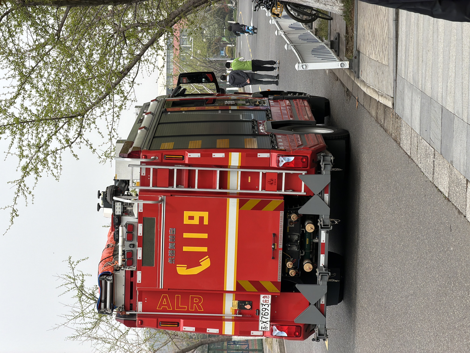 The image shows the rear of a red fire truck parked on a grey asphalt road. The number "119" is prominently displayed in yellow on the back of the truck, along with Chinese characters. Below "119" is "ALR" in red lettering. The license plate reads "苏-X7693应急".
There are people standing on the sidewalk next to the road. A bicycle is parked near the people.
The image may be in Jiangning District, China. The full phrase "广东永强奥林" is visible on the vehicle. According to the search results,广东永强奥林 is a brand of fire truck manufactured in China.