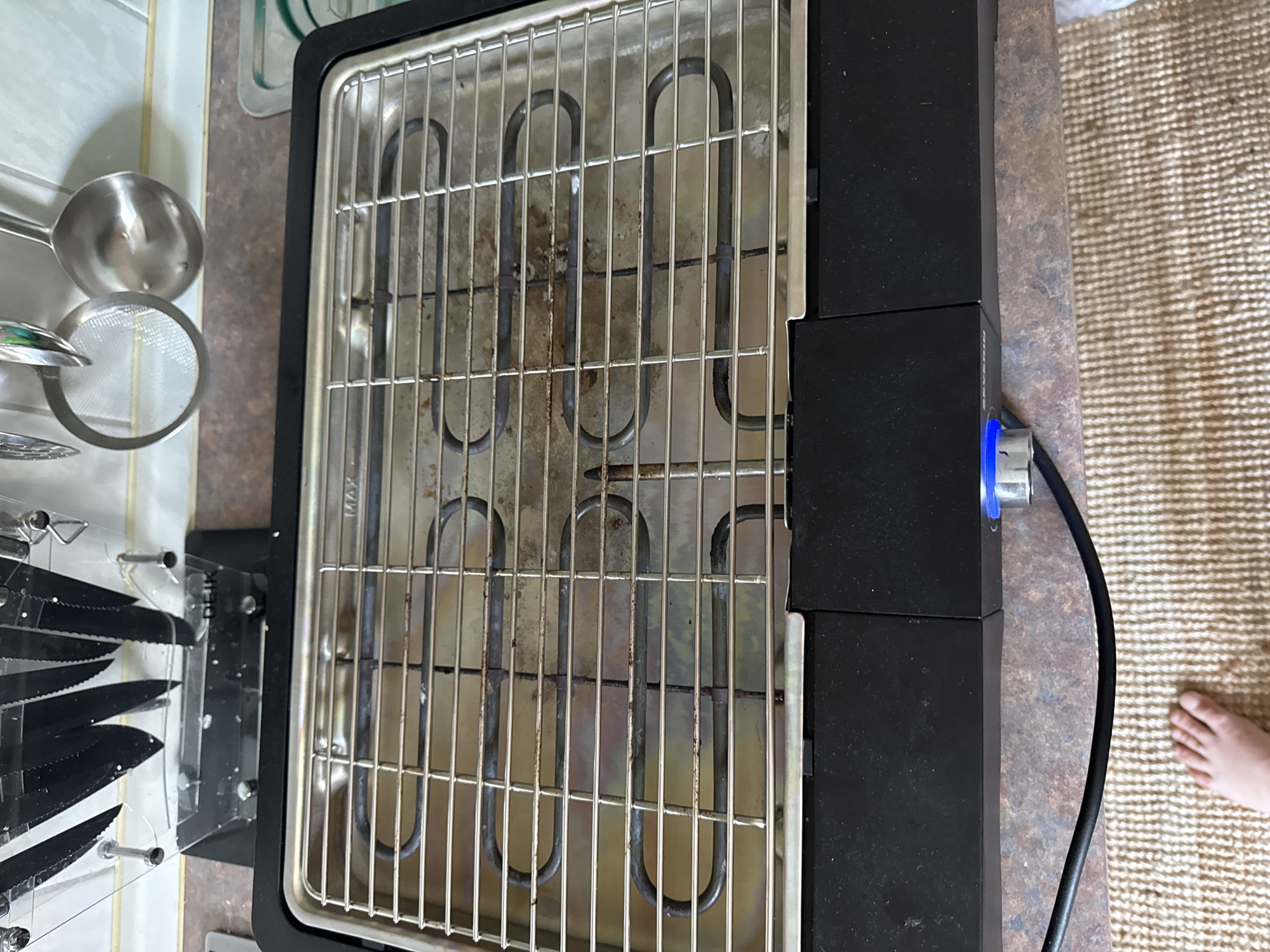 An electric grill sits on a countertop in a kitchen. The grill has a chrome grate over a heating element that is U-shaped. The grill has a black base with a blue light and a silver knob on the front. In the background, a knife block filled with black knives and kitchen utensils hang on the wall. To the right of the grill is a metal tray with a green design. A black power cord for the grill extends to the left and rests on a woven rug. A person's bare foot is visible on the rug in the lower left corner of the image. The countertop has a mottled brown and gray pattern. The wall behind the countertop is tiled with white and brown tiles. The kitchen appears to be in a residential setting. This scene is not specifically located in Höhenkirchen-Siegertsbrunn, Germany, as there are no identifying landmarks or specific indicators of this location.