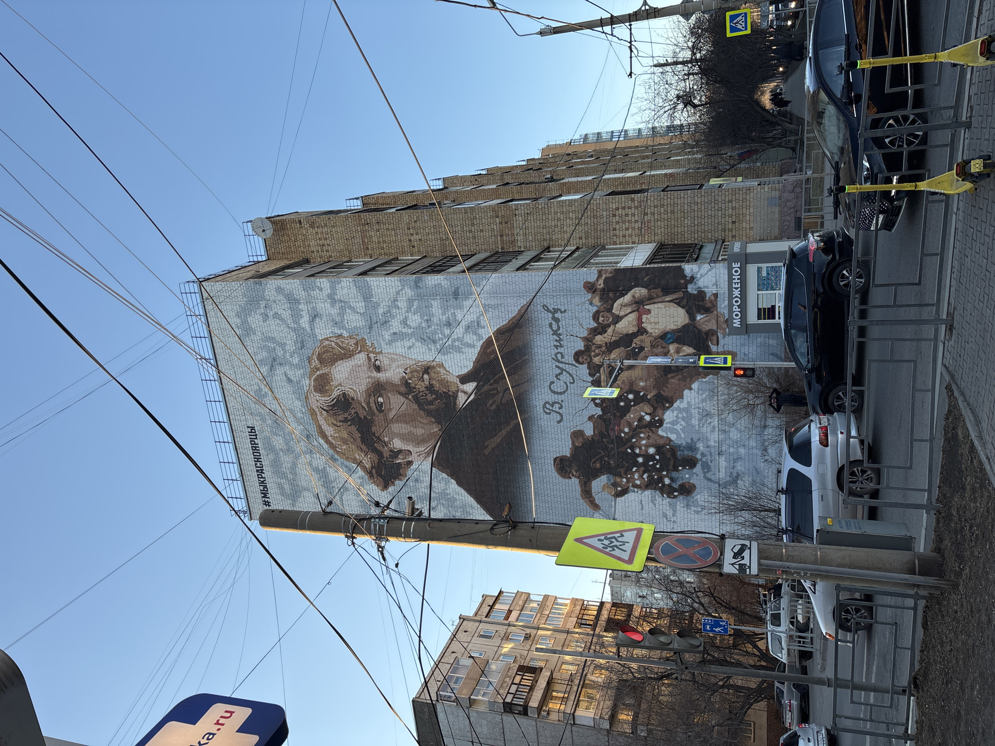 The image shows a mural depicting the Russian painter Vasily Surikov on the side of a building in Krasnoyarsk, Russia. The mural includes a portrait of Surikov and an illustration of his painting "Boyarynya Morozova". The building appears to be a multi-story apartment complex constructed of brick. Utility poles with numerous wires intersect the view. Street signs and traffic lights are visible, as are parked cars and pedestrian barriers. The environment appears to be an urban street with buildings on both sides and a clear sky above.