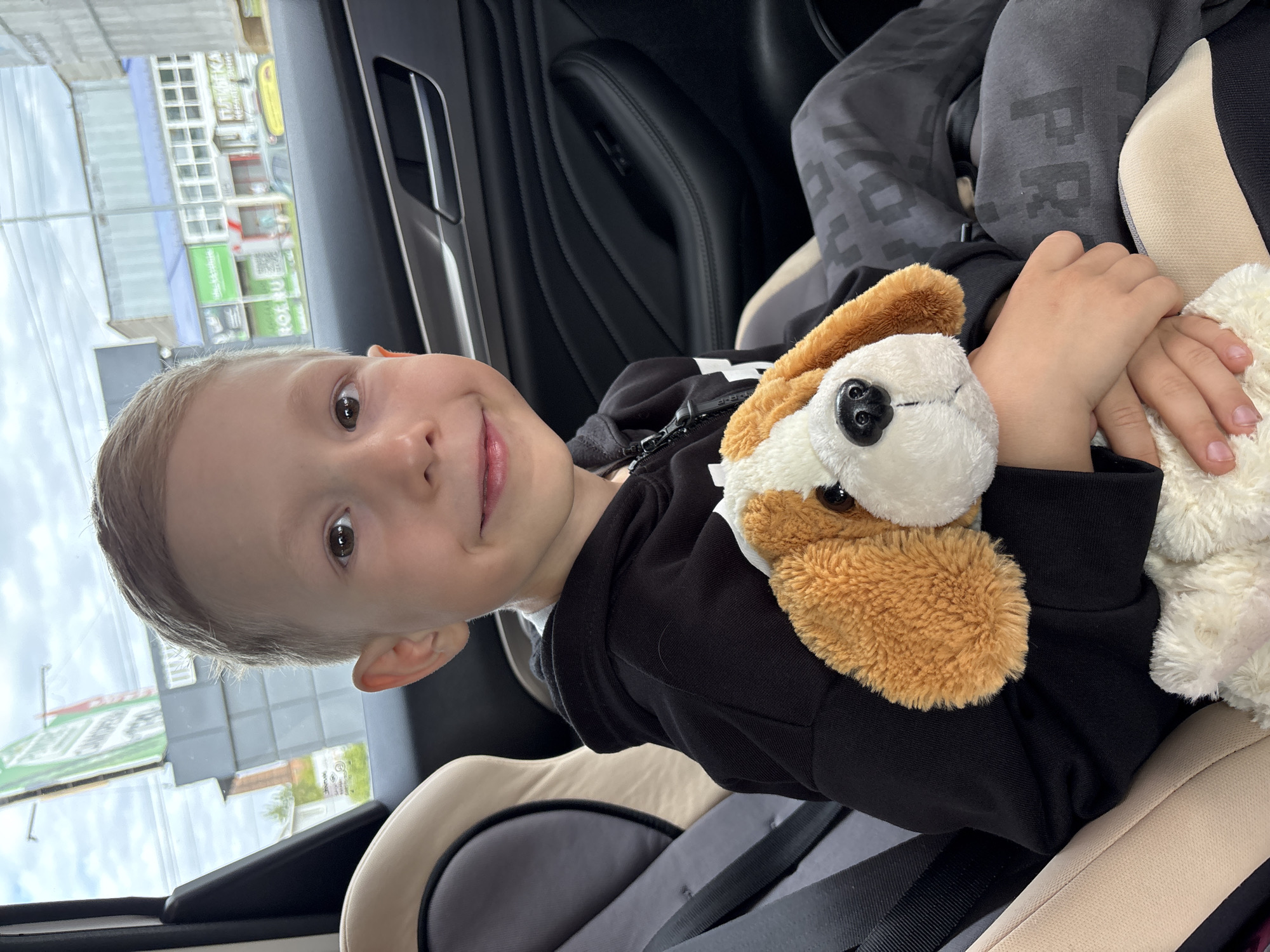 A young boy with short blond hair and brown eyes is sitting in a car seat, holding a stuffed dog. He is wearing a black hooded sweatshirt and grey sweatpants with a pixelated pattern. The car is parked outdoors, with buildings visible through the windows. The sky is cloudy. The location is the city of Chaikovsky, Russia, as indicated by signage visible in the background.