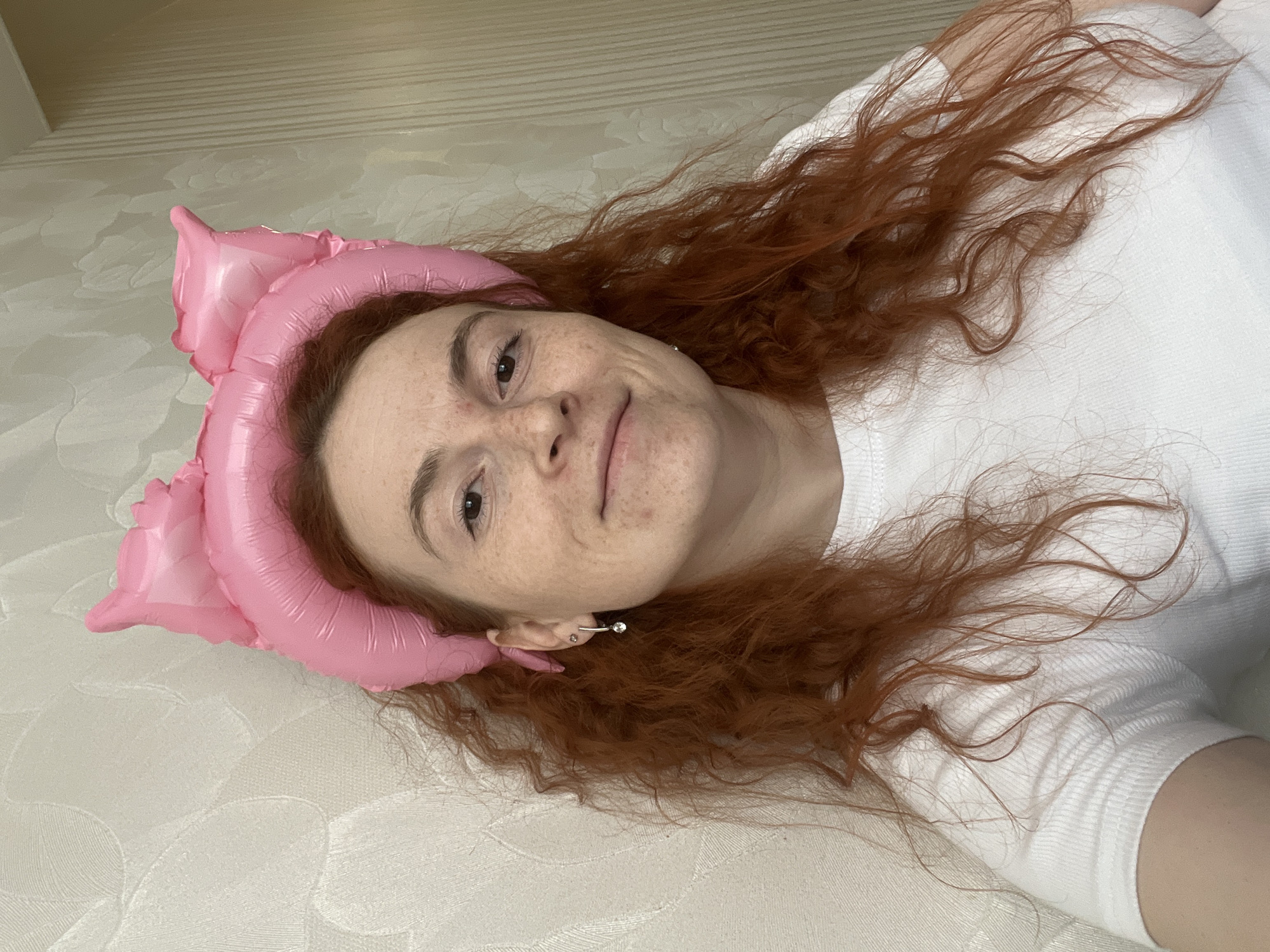 A light-skinned woman with freckles and red curly hair lies on a cream-colored textured surface, presumably a bed. She is wearing a white shirt. A pink, inflatable object that resembles a flower or a cat’s head rests near her head. She is smiling and looking directly at the camera. Her left ear is visible, with a piercing. The location is the city of Chaikovsky, Russia.