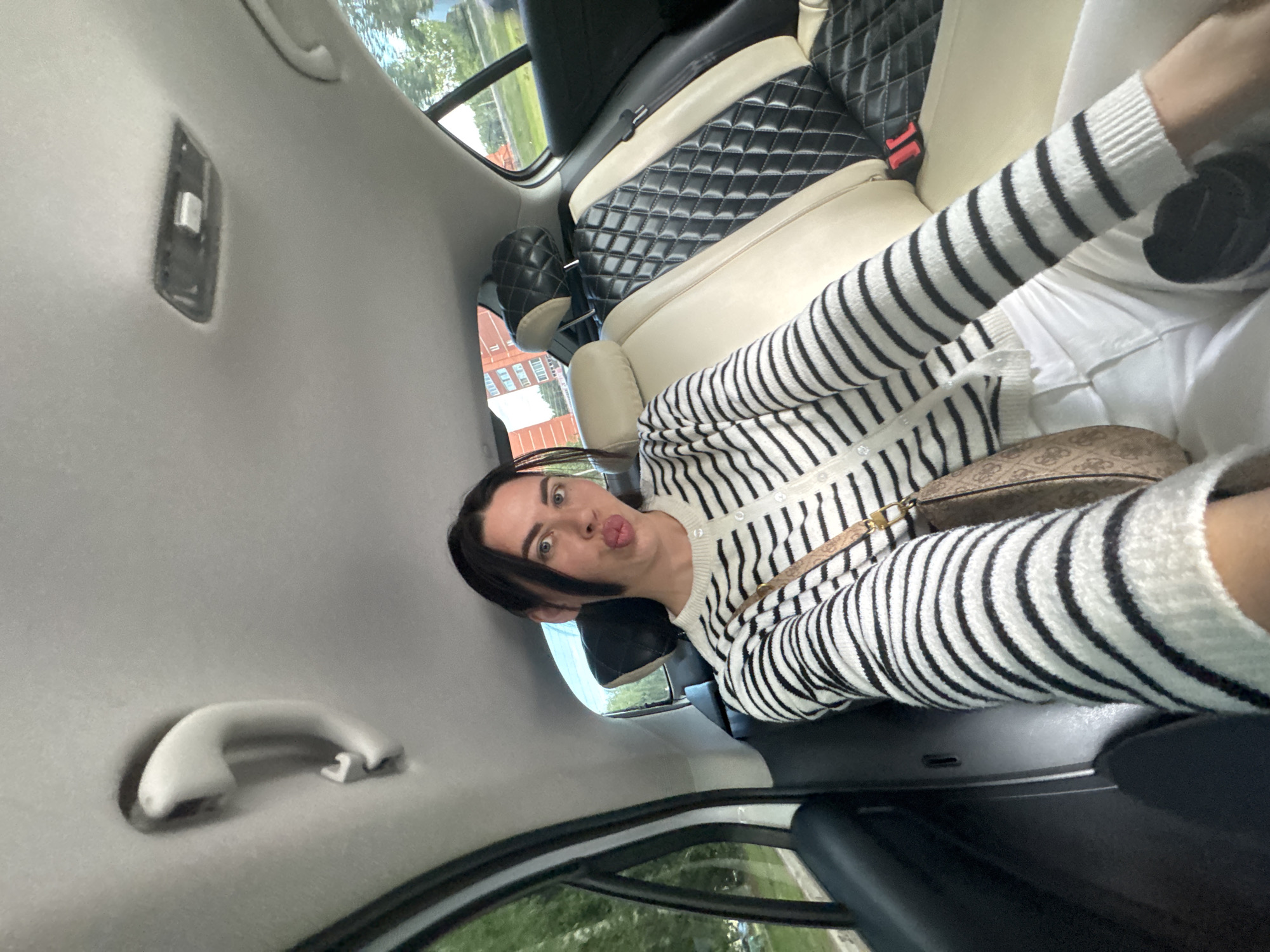 A person is sitting in the backseat of a car, facing the camera with their mouth pursed. They are wearing a black and white striped cardigan and white pants, and a patterned purse is slung across their chest. The car's interior is visible, with beige and black diamond-stitched seats. The person's left arm is extended, holding the phone that captured the image. The background outside the car window shows trees and buildings, suggesting an urban or suburban setting. The image was taken in Chaikovsky, Russia, indicated by the geotag.
