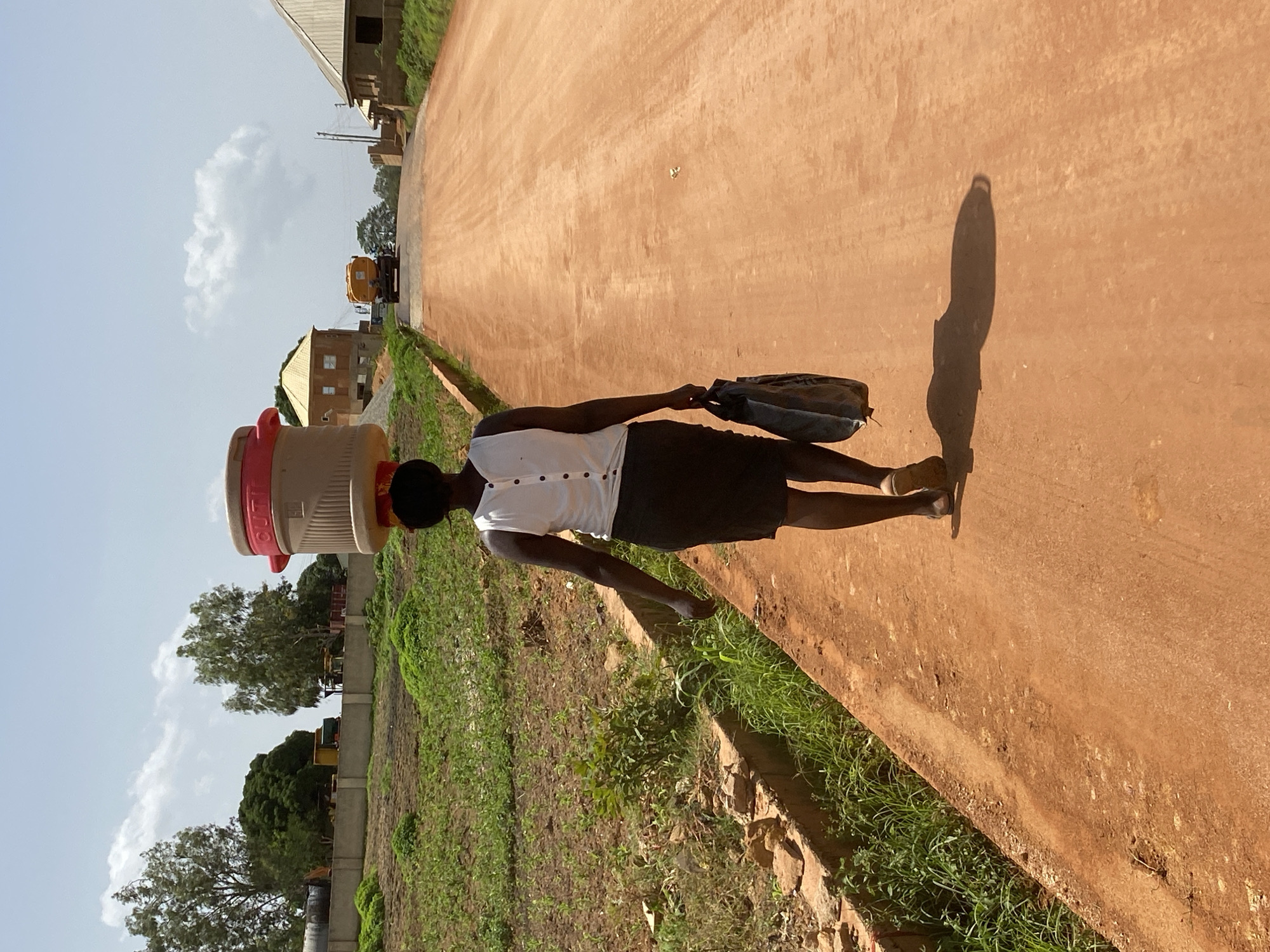 A dark-skinned woman walks away from the camera on a dirt road in Bukuru, Nigeria. She is carrying a large, beige and red water cooler on her head and a dark blue bag in her right hand. She is wearing a white sleeveless top with buttons down the back and a dark skirt. To the left of the road, there is a ditch with some vegetation and scattered rocks. Further back on the left, there is a wall with some trees and construction equipment. On the right side of the road, there are houses with yellow roofs and some construction. In the distance, a yellow truck is visible on the road. The sky is light blue with some white clouds.