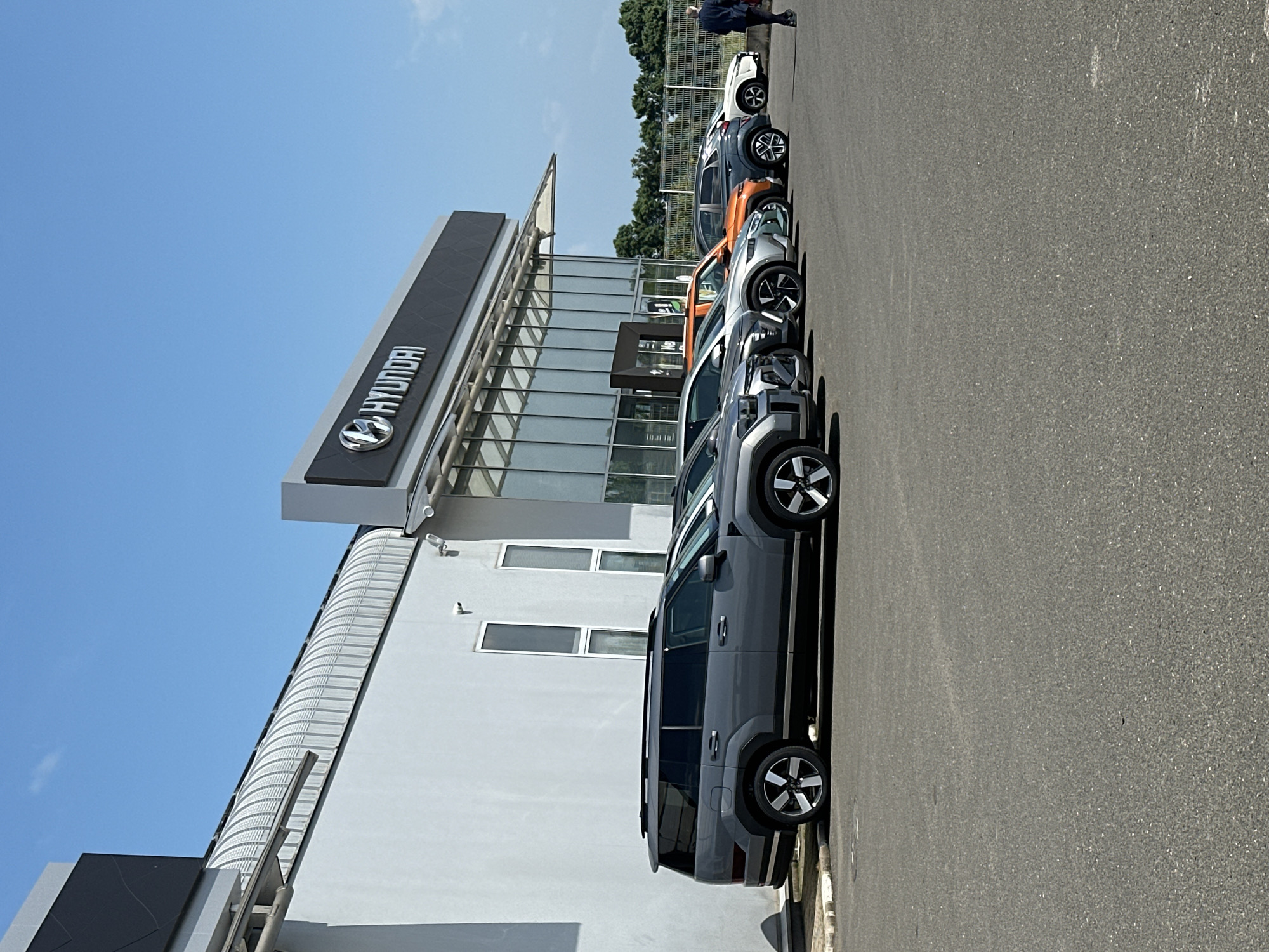 The image, when viewed in its correct orientation, displays the exterior of a Hyundai car dealership on a clear, sunny day. The main subject is a modern, white building with large glass panels and dark trim, which serves as the dealership. A prominent black sign featuring the silver "HYUNDAI" logo and text is affixed to the upper facade of the building.
Parked in a neat line on a grey asphalt lot in front of the dealership are several new Hyundai vehicles, predominantly SUVs or crossovers. These cars are presented in various colors, including dark grey, light grey, orange, and white, each showcasing distinct wheel designs.
The ground consists of a textured grey asphalt surface that covers the foreground and extends towards the background. Above the building and vehicles, a bright, clear blue sky with minimal cloud cover is visible, indicating pleasant weather.
In the upper right section of the image, partially obscured by a fence and some trees, a single person is seen. They are dressed in a dark jacket and light-colored trousers, standing near the far end of the parked cars. Their activity is not clear, but they appear to be either walking or observing the vehicles. The strong lighting suggests it is daytime, possibly midday or early afternoon, due to the shadows cast by the cars and building.